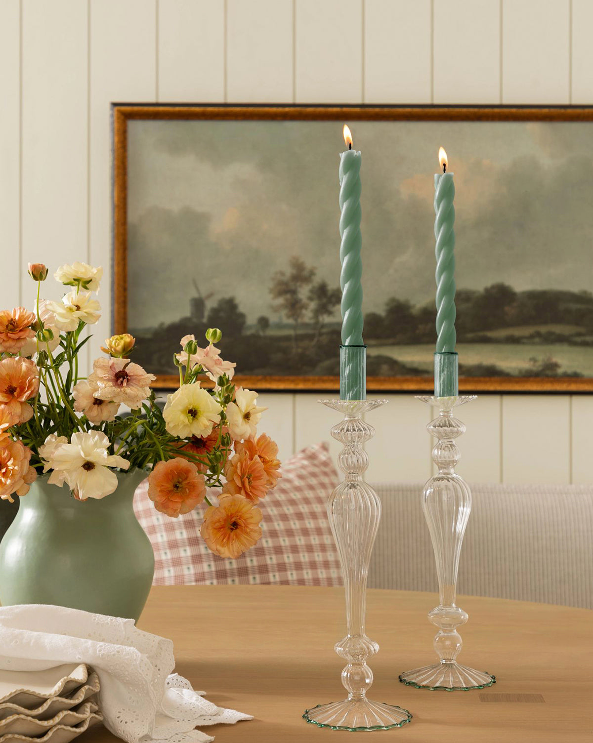 A vase of peach and cream flowers sits on a wooden table beside two lit, twisted blue candles in a McGee & Co. x Loeffler Randall Effie Taper Holder. A folded white napkin is nearby, with a landscape painting visible in the background by McGee & Co.