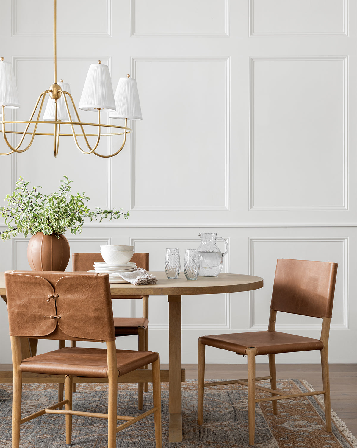 A modern dining area features a round wooden table, four Danson Dining Chairs with brown leather seats, tableware, a potted plant, and a gold chandelier against a white paneled wall for a minimalist look, by McGee & Co.