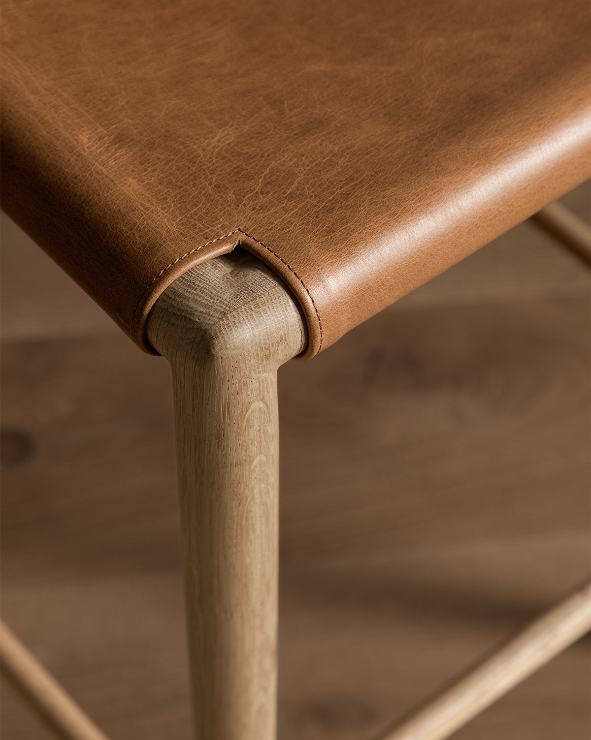 A close-up of the Danson Counter Stool corner shows its smooth leather seat with visible stitching, neatly wrapped over a rounded light wood frame. The stool’s minimalist design pairs seamlessly with a similarly toned wooden floor in the background, by McGee & Co.