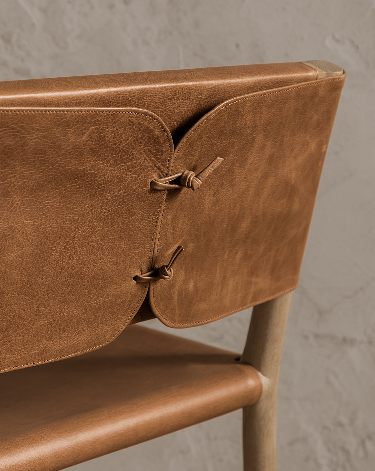 Close-up of the Danson Counter Stool, showcasing its light brown leather seat and backrest with two panels connected by leather cords, highlighting minimalist design against a light textured wall, by McGee & Co.