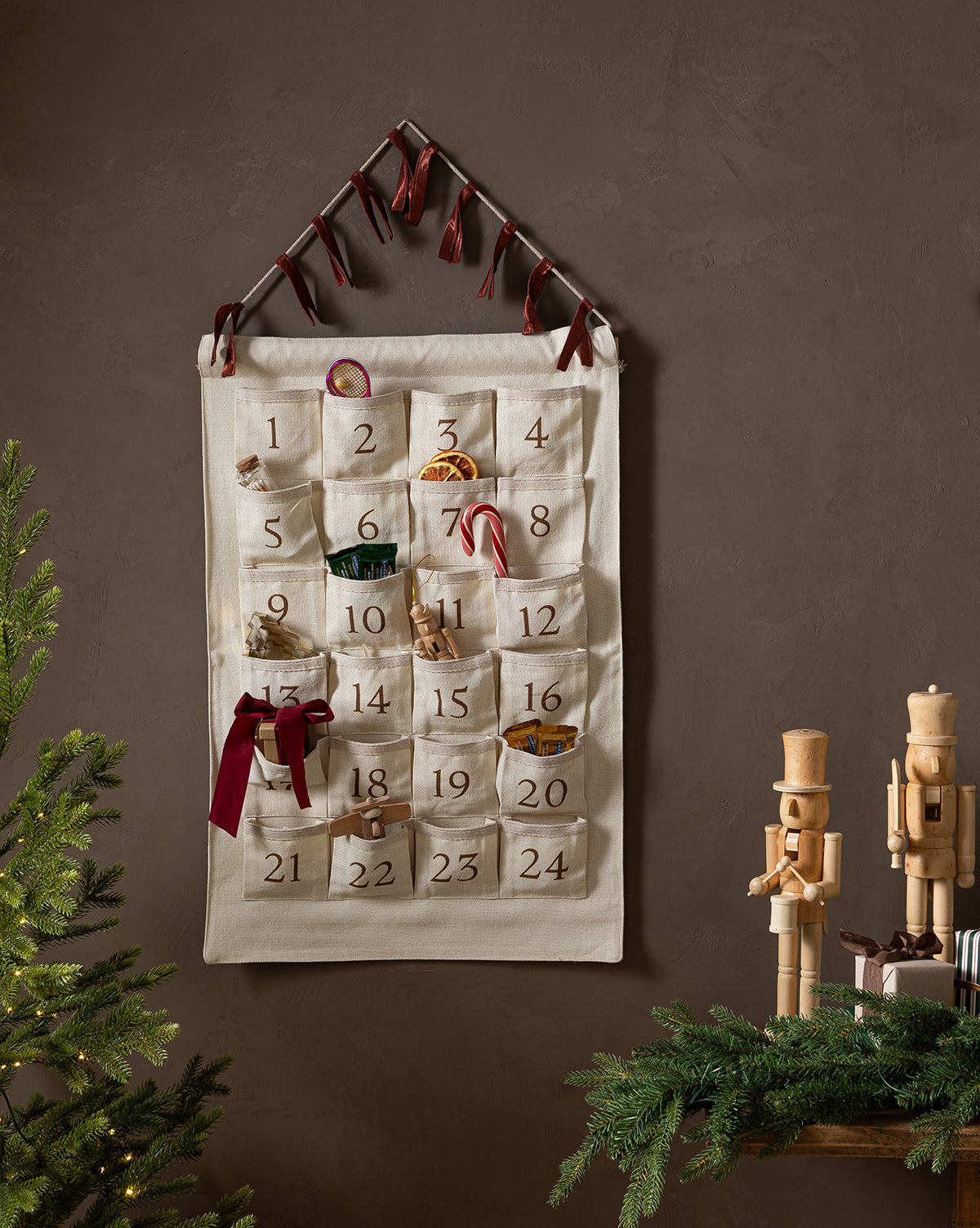 The Advent Calendar, made of cotton linen with numbered pockets for small gifts and ribbons, hangs on the wall. Below, two wooden nutcrackers and pine branches sit near a Christmas tree, creating a charming holiday scene, by McGee & Co.