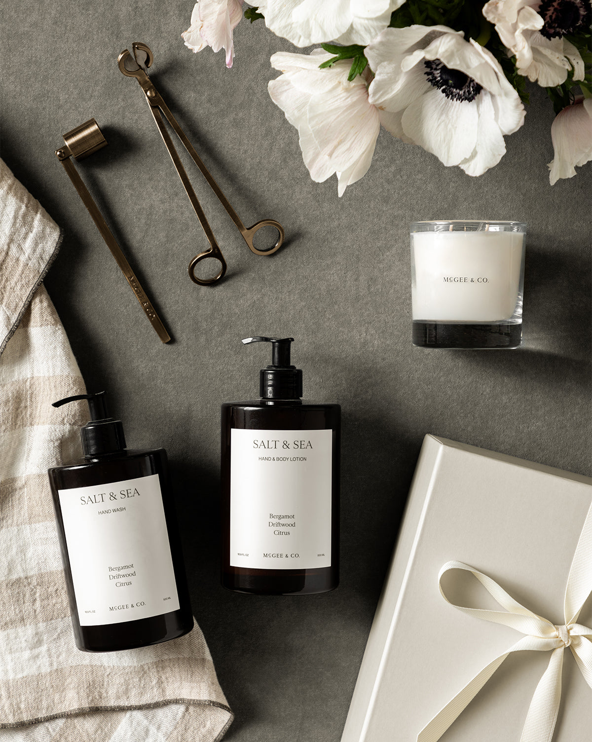 Flat lay of luxury self-care items on a gray surface, featuring the Salt & Sea Gift Set with lotion, hand wash, signature scented candle, candle accessories, striped towel, white ribboned gift box, and white flowers in the corner, by McGee & Co.
