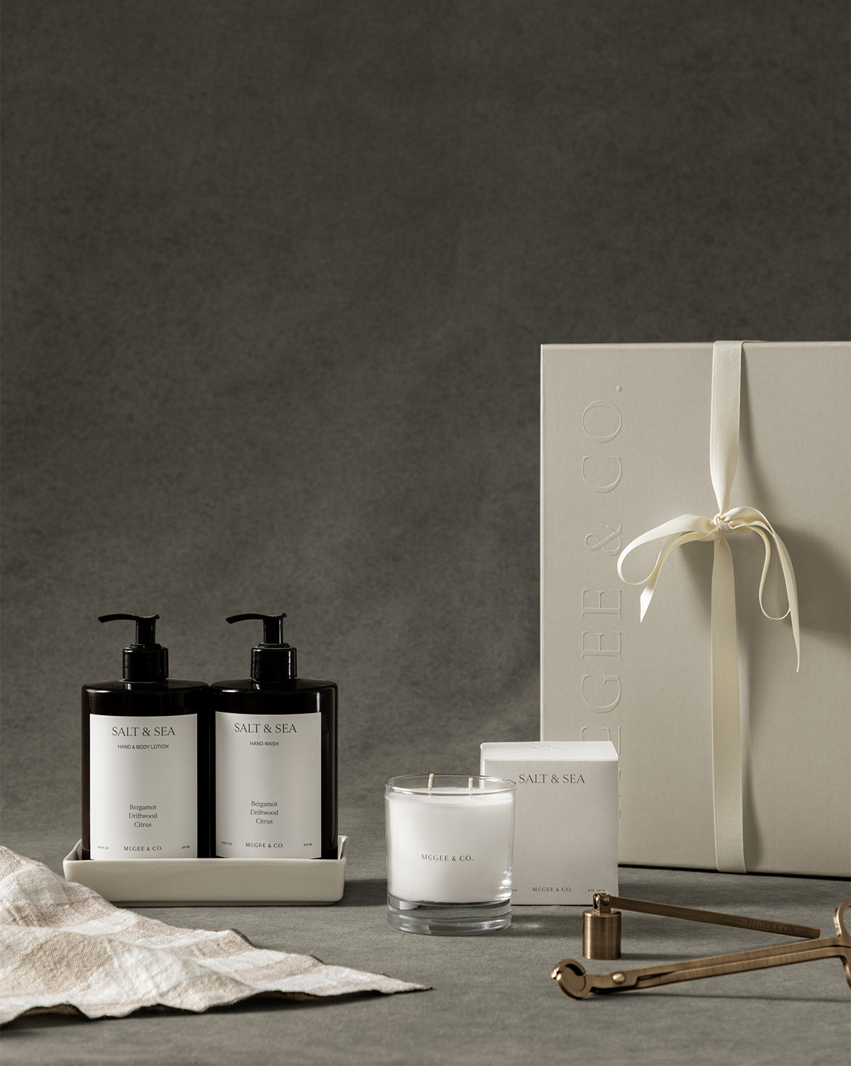 The Salt & Sea Gift Set features two pump bottles, a candle in a glass jar with box, tray, wick trimmer, and an elegant gift box tied with cream ribbon—all displayed on a neutral background by McGee & Co.