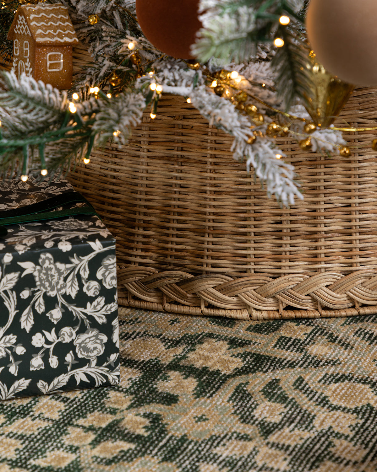 A close-up of a holiday tree with frosted branches, ornaments, string lights, and a wrapped gift on a patterned rug. The tree sits in the Braided Wicker Collar for a cozy festive touch, by McGee & Co.