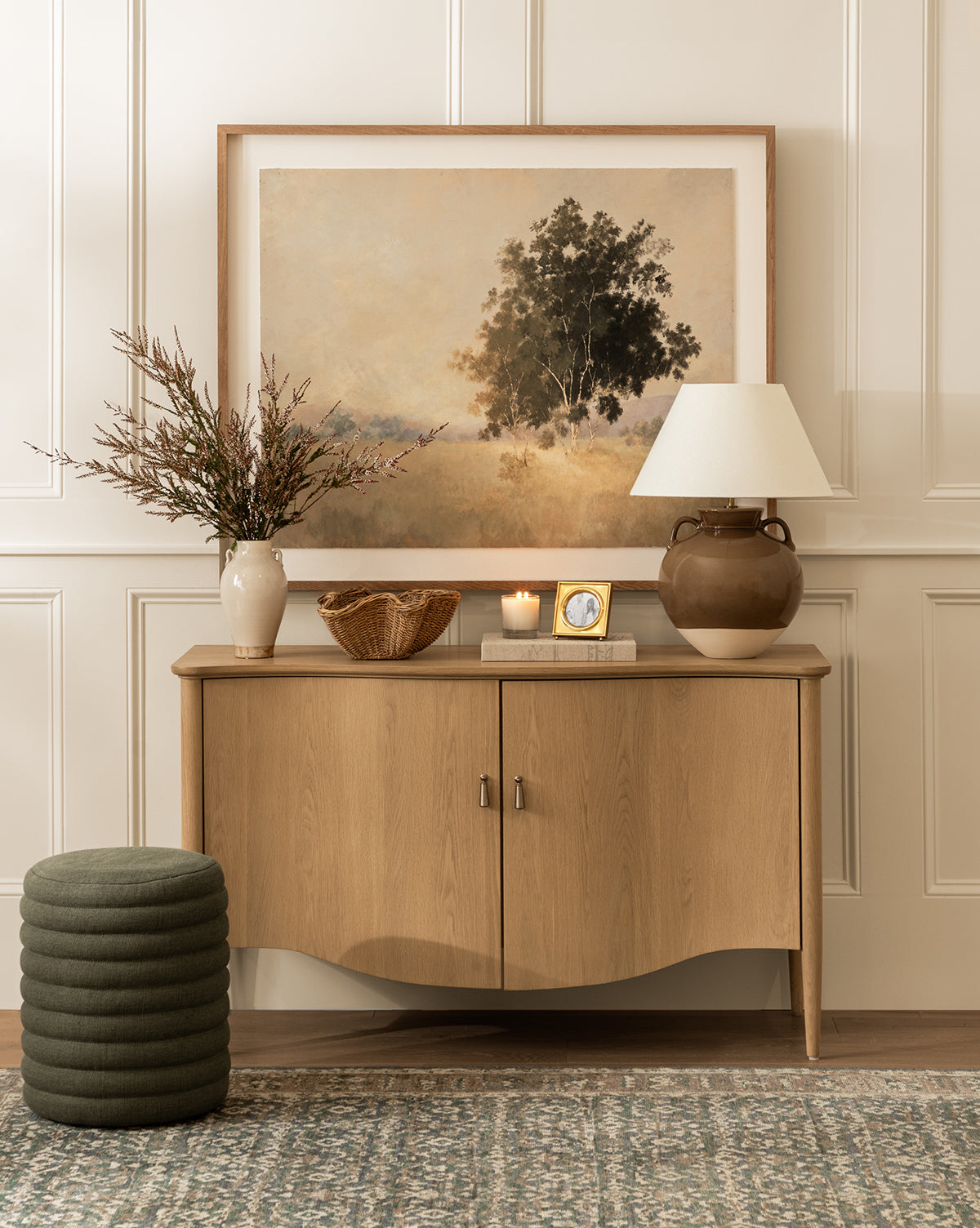 A wooden sideboard topped with a lamp, vase of branches, basket, candle, clock, and landscape painting sits against a paneled wall. In front are a green pouf and the Made Goods Jefferson Stool on a patterned rug, all curated by McGee & Co.