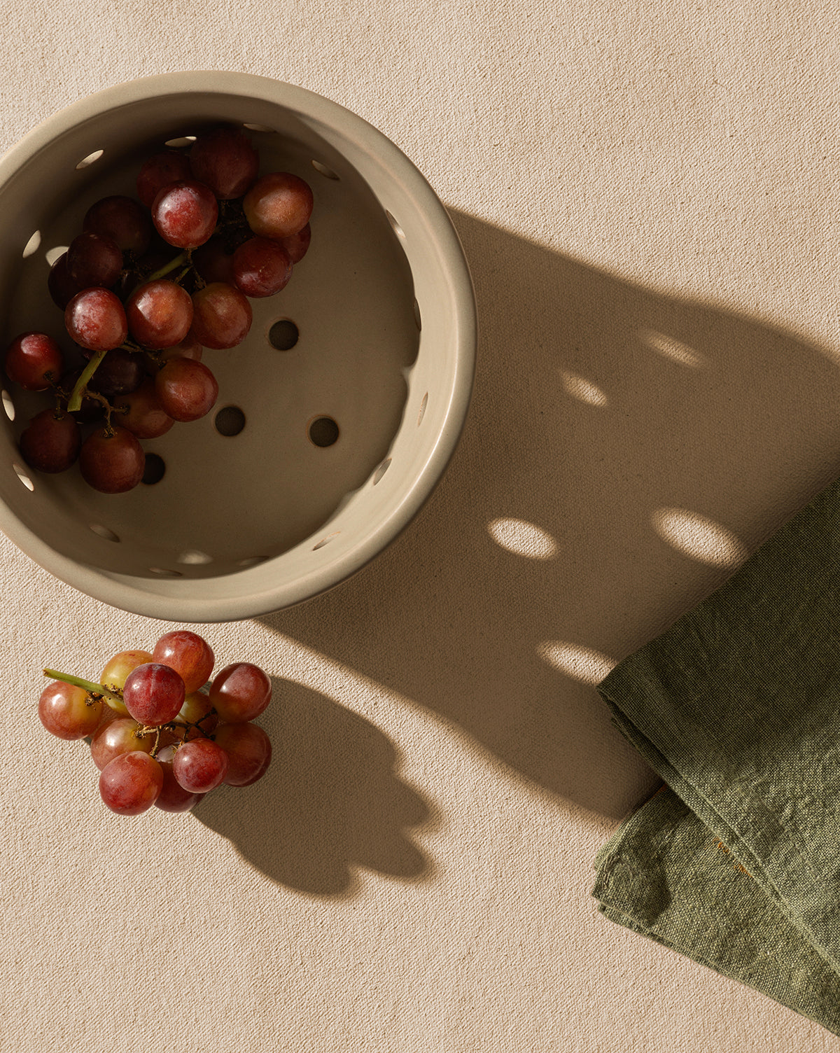 A beige Coen Berry Bowl with a matte finish holds red grapes on a light textured surface. A small bunch sits beside the stoneware bowl, near a folded green napkin. Both the Coen Berry Bowl and grapes cast distinct shadows by McGee & Co.