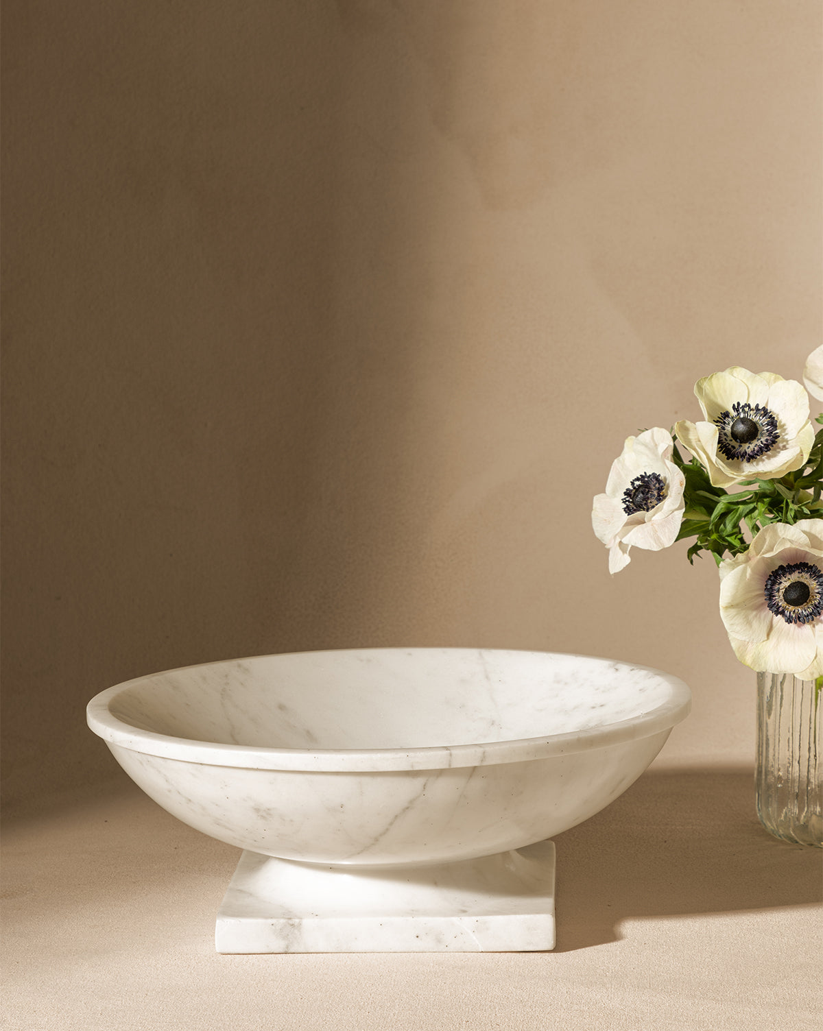 The Tayva Bowl, a sculptural white marble piece with a square base, rests on a neutral surface beside a glass vase of white anemones with dark centers against a beige background, by McGee & Co.