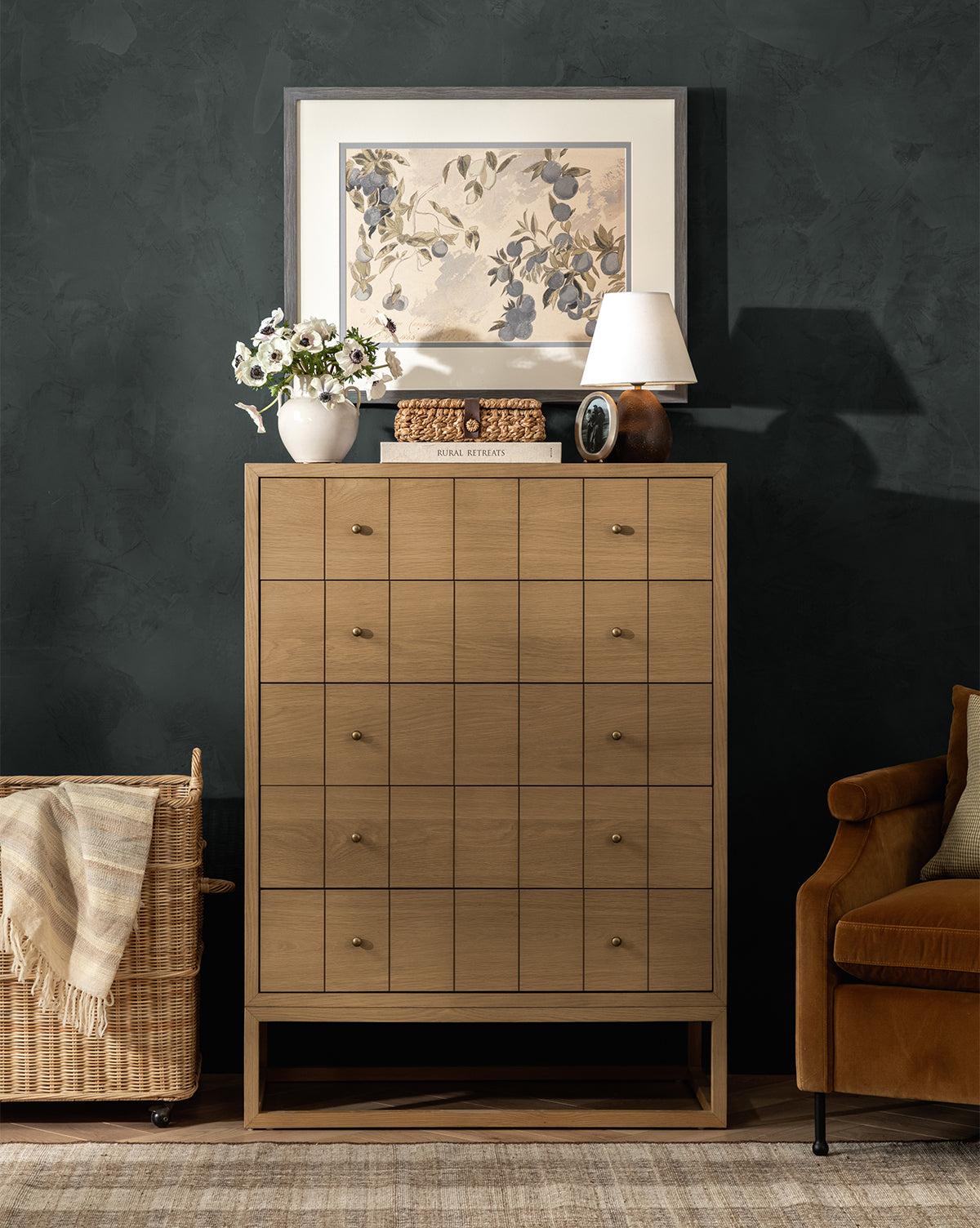 A wooden dresser with several drawers stands against a dark wall, topped with the Vedruna Table Lamp and decor. Above hangs a floral art print; nearby are a wicker basket and brown armchair, by McGee & Co.