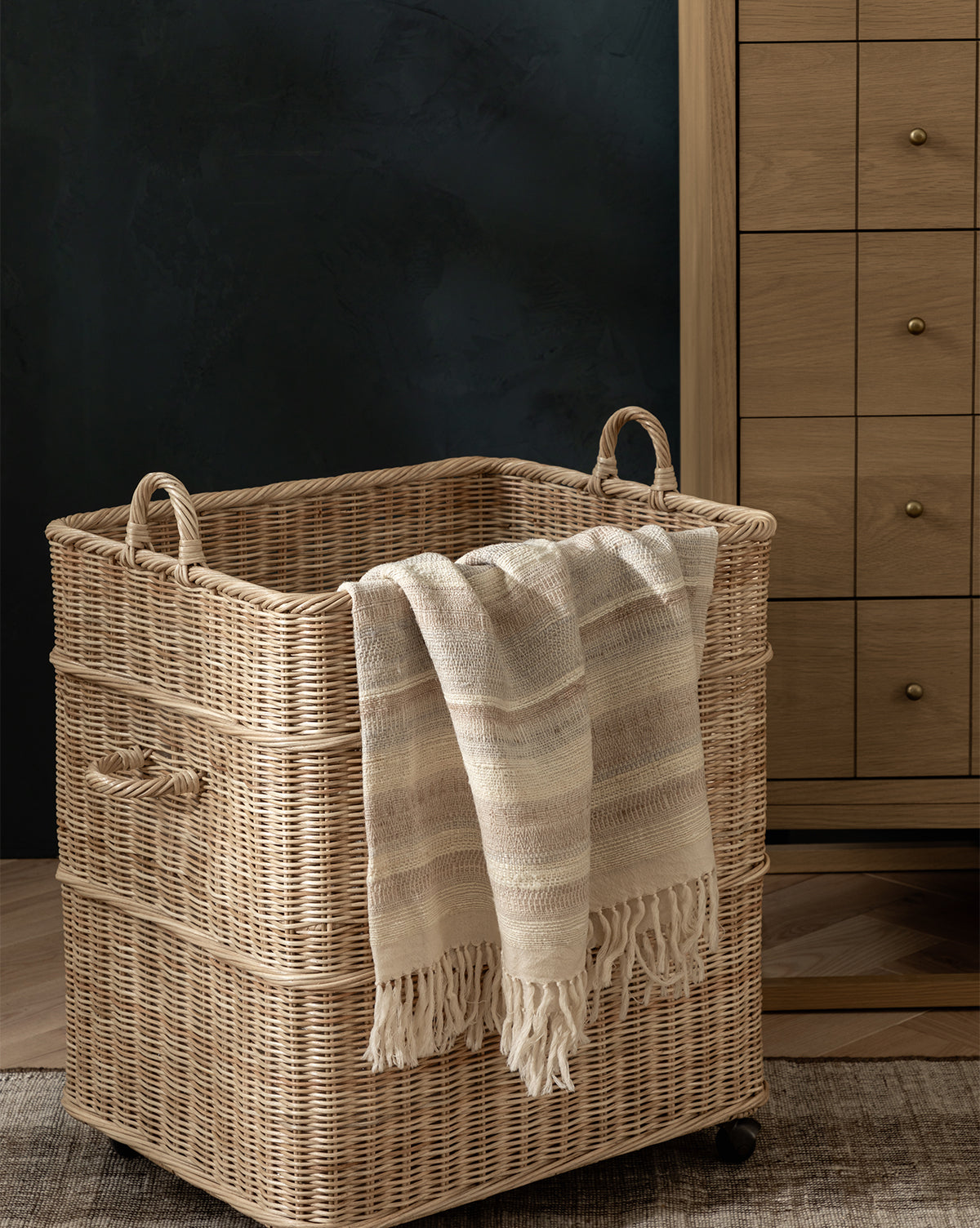 The Chiara Laundry Basket with natural wicker finish, handles, and wheels sits on a wooden floor. A cream fringed blanket hangs over it, while a wooden dresser with brass knobs completes this cozy laundry room scene, by McGee & Co.