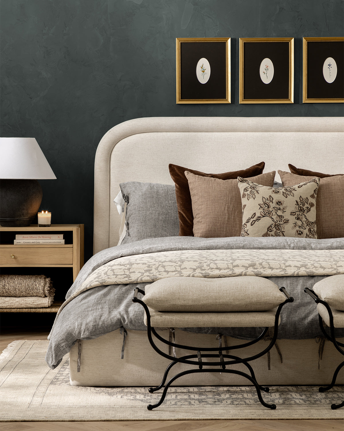 A stylish bedroom featuring a beige upholstered bed, neutral bedding with the Floral Matelasse Coverlet, layered pillows, a wooden nightstand with lamp and candle, two cushioned benches, and three framed botanical prints on a dark green wall.
