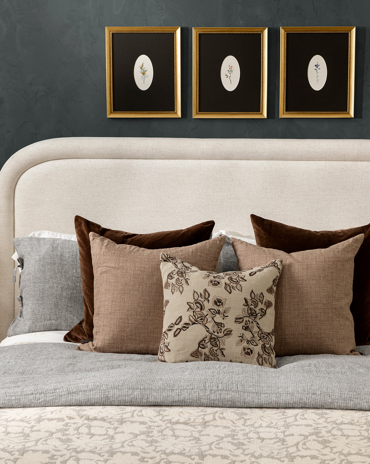 A neatly made bed with gray and beige linens, the Bellevue Velvet Pillow Cover among brown and floral pillows, plus a light upholstered headboard. Three framed botanical prints hang on a dark wall above the bed, by McGee & Co.