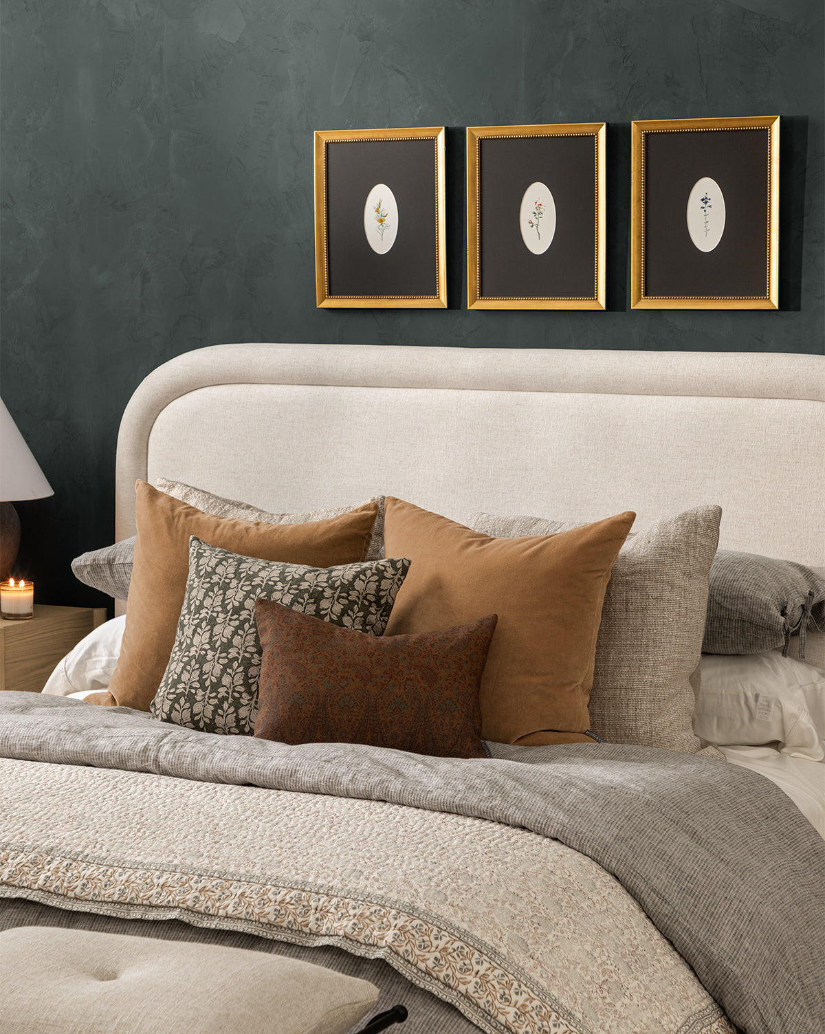 A cozy bedroom with a beige upholstered headboard, layered neutral bedding, assorted throw pillows—including a cotton pillow and the DUPE Bellevue Velvet Pillow Cover—and three botanical prints on a dark green wall above the bed, plus a lamp and candle on the bedside table, by McGee & Co.