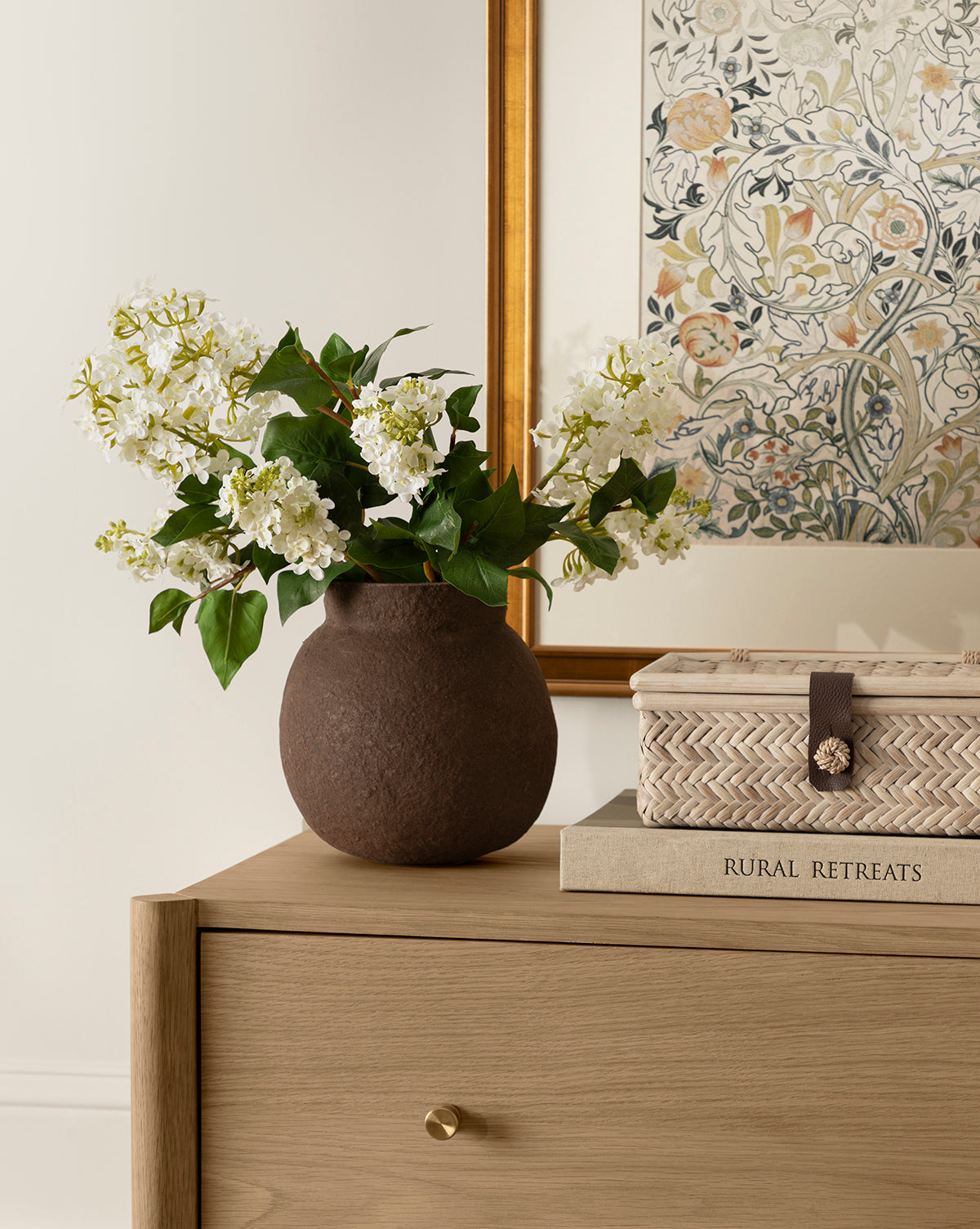 The Frederick Paper Mache Vase with white flowers sits on a light wooden dresser beside a woven box and the book Rural Retreats, while a floral-framed artwork hangs on the wall in the background, by McGee & Co.