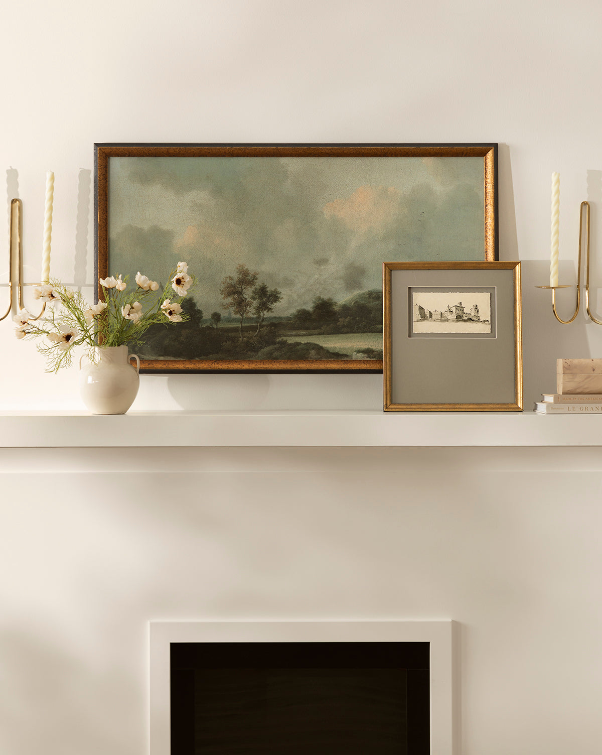 A mantel styled with a vase of white flowers, two Sepia City gallery artworks depicting romantic town scenes, two twisted candle wall holders, and a stack of books set against a neutral wall.