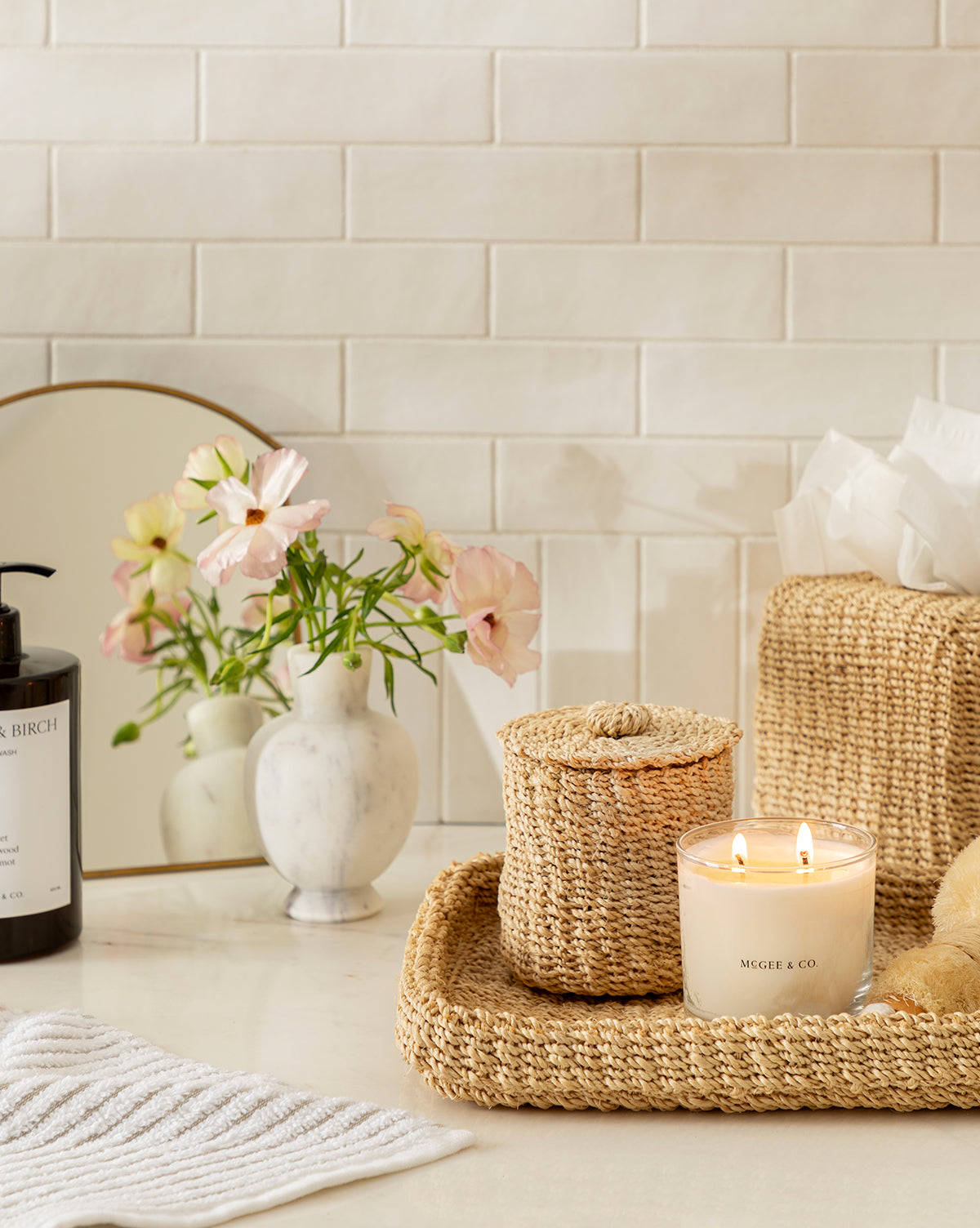 A stylish bathroom counter features a Thalia Woven Canister holding essentials next to a lit candle, tissue box, marble vase of pink flowers, and hand soap, creating an organized and fresh look on the white countertop.