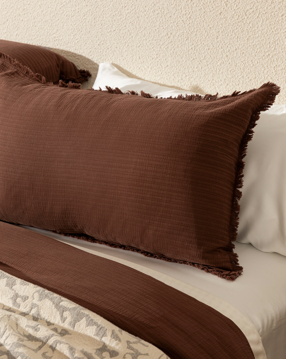 A close-up of a neatly made bed featuring Hendricks Chocolate Cotton Shams (Set of 2) with fringe edges, a white 100% cotton bedsheet, and a matching brown blanket, set against a textured beige wall.