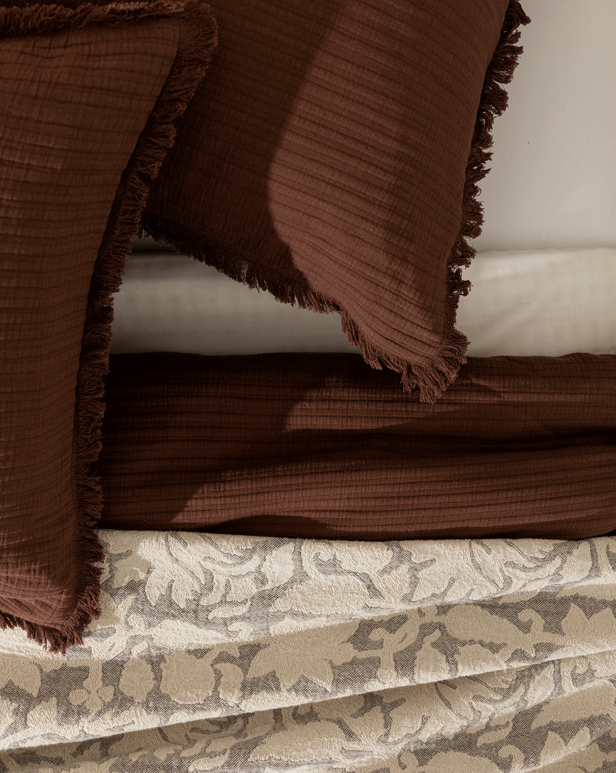 A close-up shows 100% cotton bedding with a textured brown quilt and Hendricks Chocolate Cotton Shams (Set of 2) featuring fringed edges, layered over a beige blanket with a subtle leafy pattern.