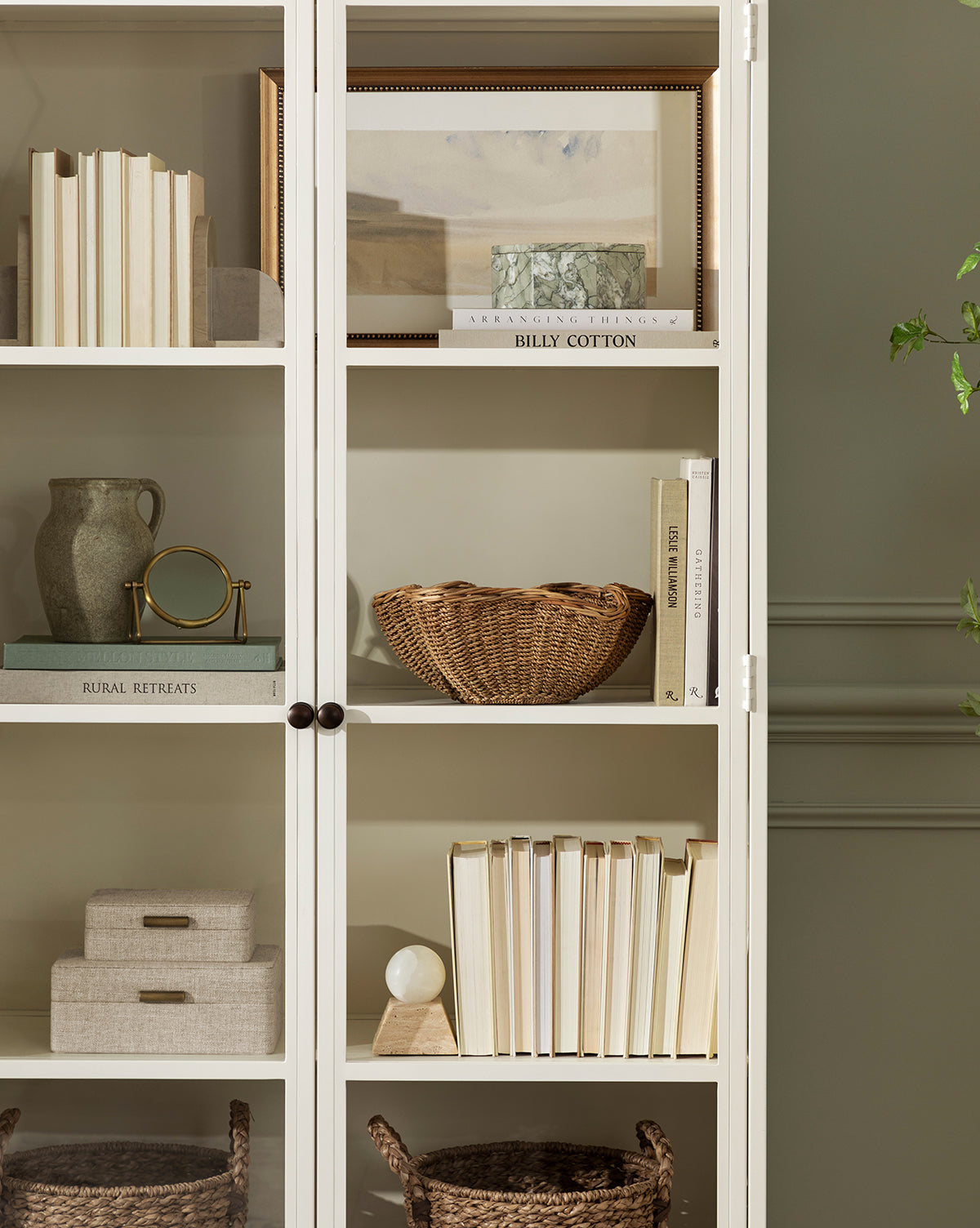 A white cabinet with glass doors displays books, decorative boxes, a framed picture, the Sabine Pitcher Vase, a gold magnifying glass, a wicker bowl, and baskets. A leafy plant is partly visible on the right, by McGee & Co.