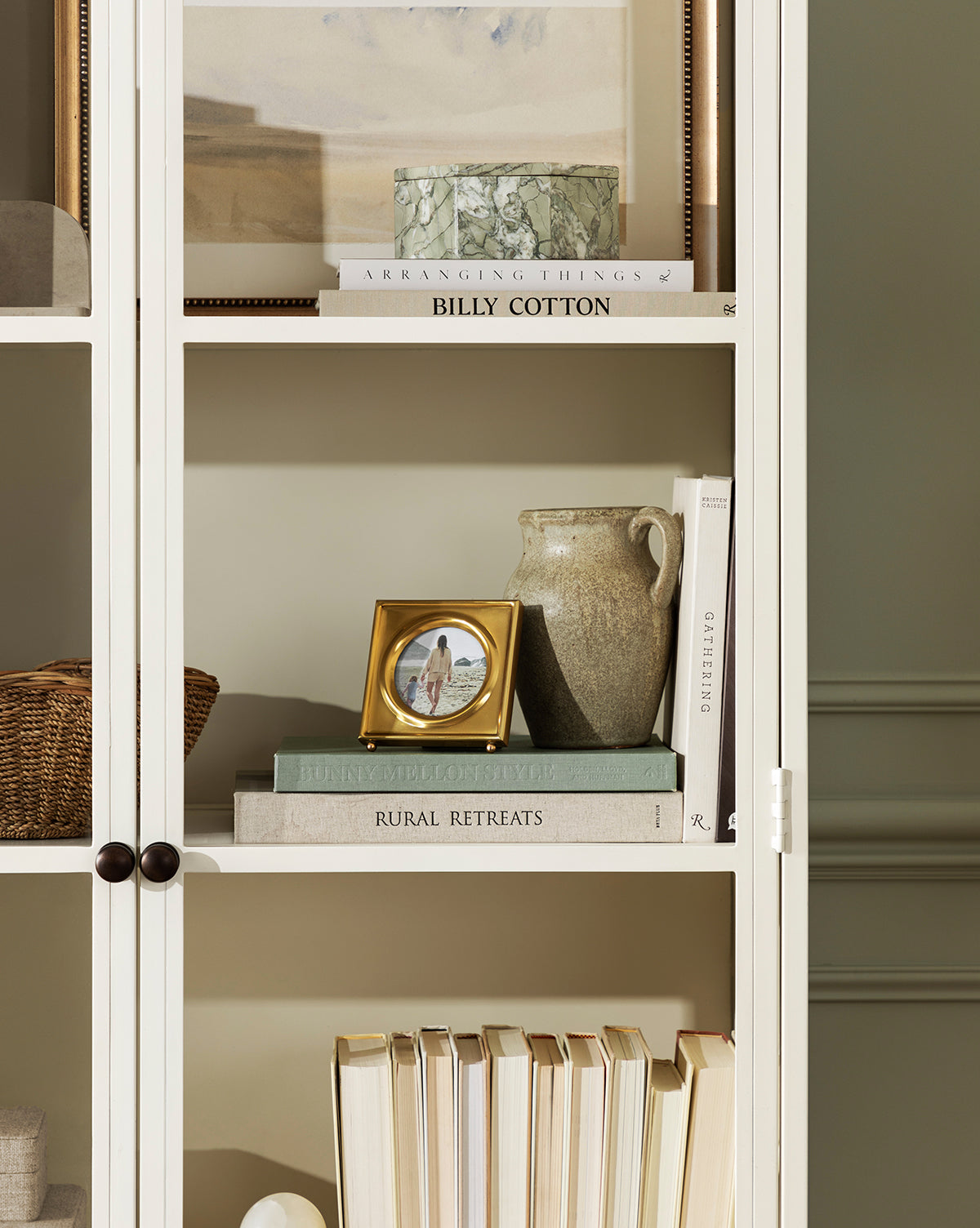 A white bookshelf with decorative items, including stacked books, a framed photo, a ceramic jug, the Paolo Marble Box, a woven basket, and neatly arranged books against a light-colored wall.