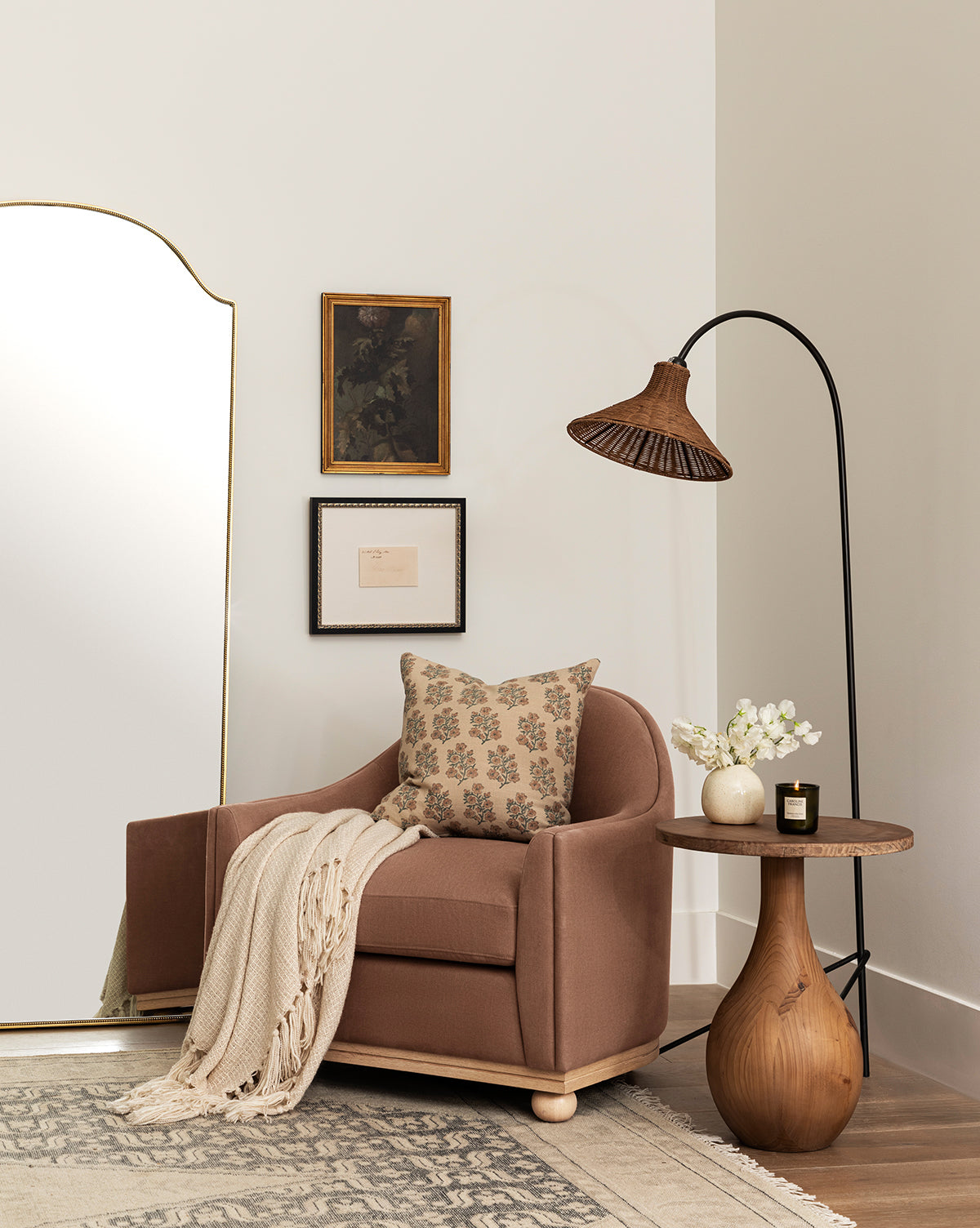A cozy reading nook with a brown Bunnell Lounge Chair (Ready to Ship), patterned pillow, and cream knit throw beside a round wooden side table topped with flowers and a candle. A floor lamp arches above and framed art decorates the white wall, by McGee & Co.