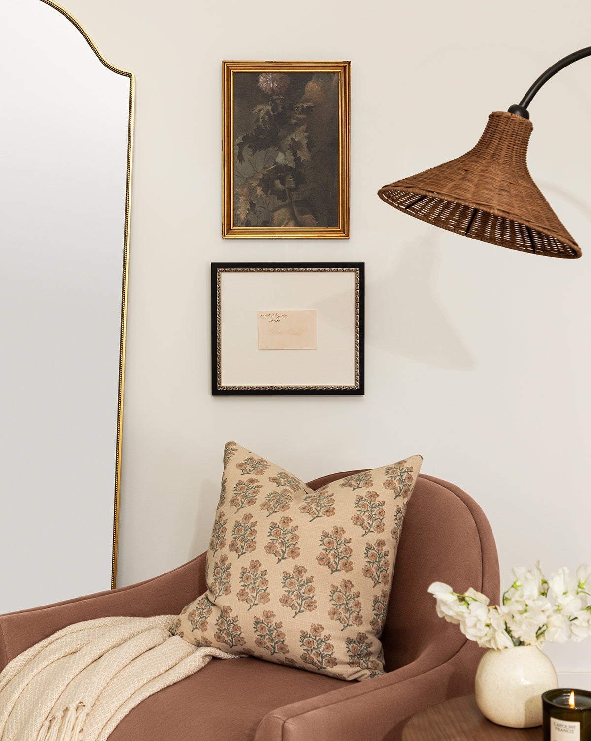A cozy corner with a mauve armchair, floral pillow, and cream throw blanket. Above hangs Thistle Down flower wall art in an antique gold frame. White flowers, a candle nearby, and a tall mirror to the left complete the space, by McGee & Co.