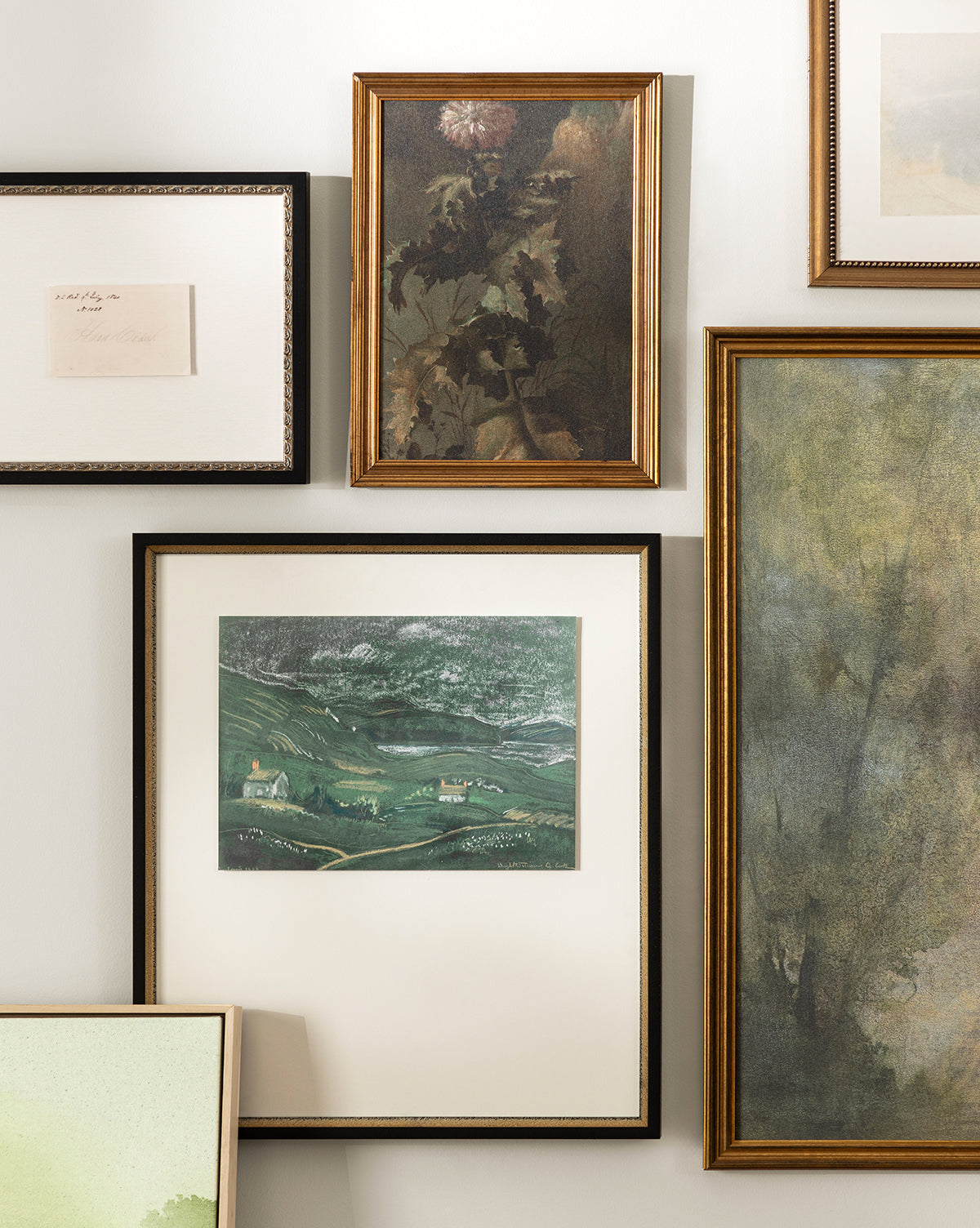 A gallery wall displays various framed artworks, including Thistle Down, an abstract painting, a green hills landscape, flower wall art in an antique gold frame, and a handwritten note—all beautifully arranged on a light wall by McGee & Co.