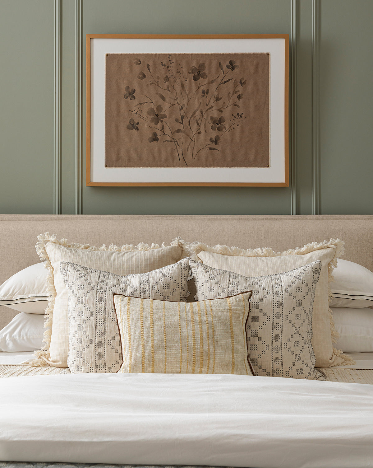A neatly made bed with cream and beige pillows, a light blanket, and a beige headboard sits under Consider the Wildflowers by Katie Mulder in a maple frame, displayed on a green-paneled wall, by McGee & Co.