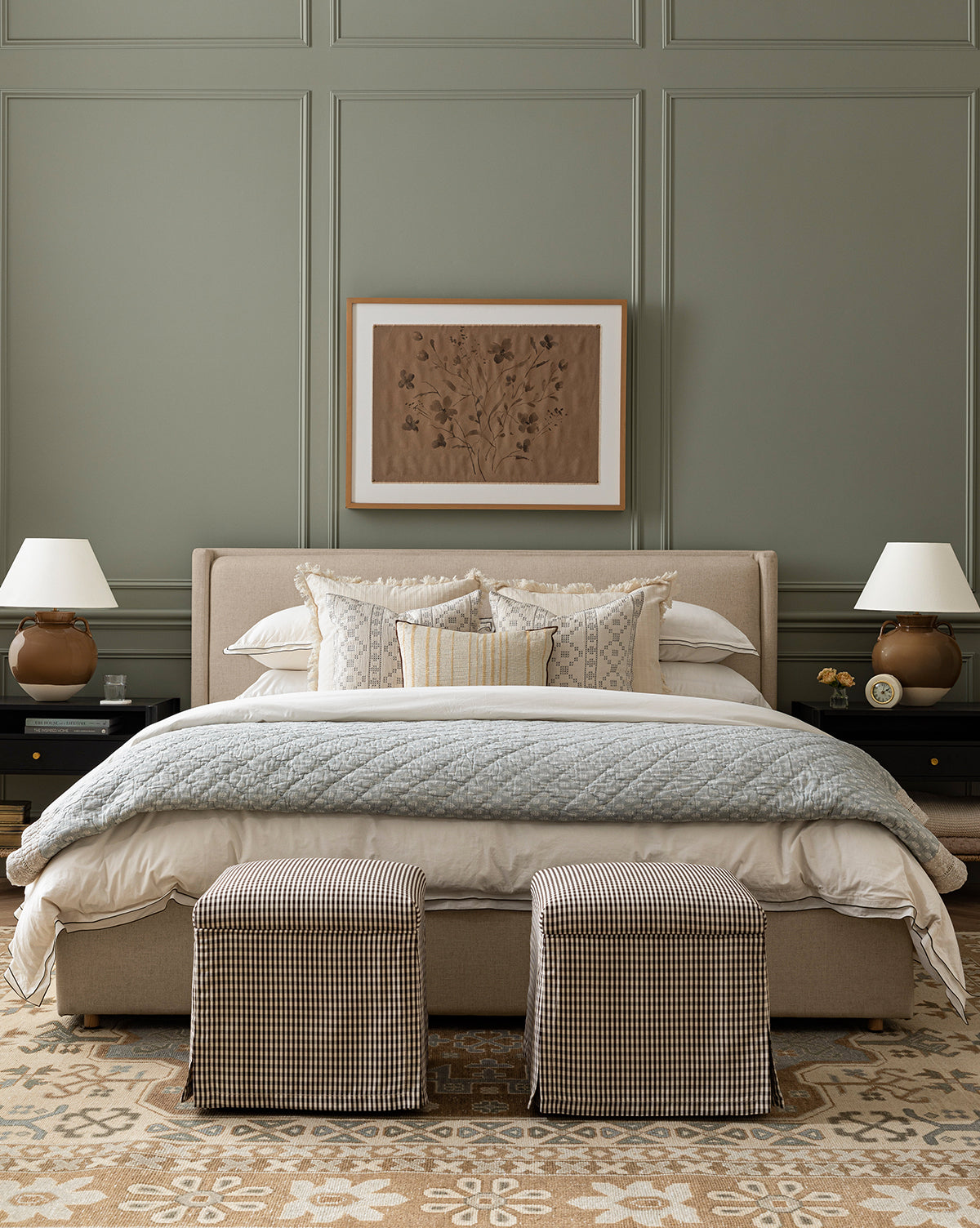 A stylish bedroom with a beige Ria Bed Low Headboard, layered white and patterned pillows, a quilted blanket, two checkered stools at the foot, matching bedside tables with lamps, and framed artwork on a green-paneled wall by McGee & Co.