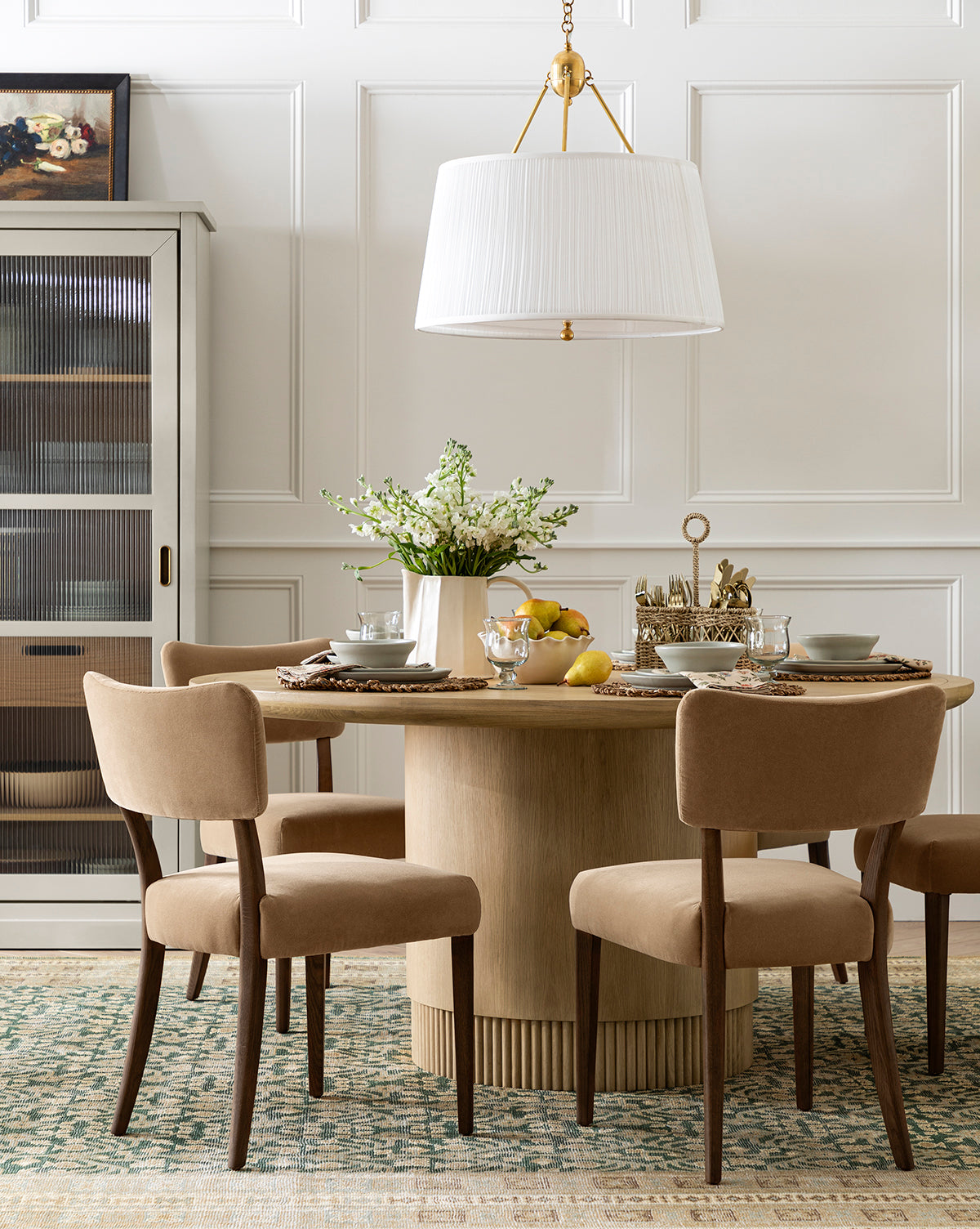 A dining room with a Gilroy Round Oak Dining Table with a reeded base, four beige upholstered chairs, a large white pendant light, elegant tableware, a patterned rug, and cabinet create this neutral-toned heritage oak finish setting by McGee & Co.