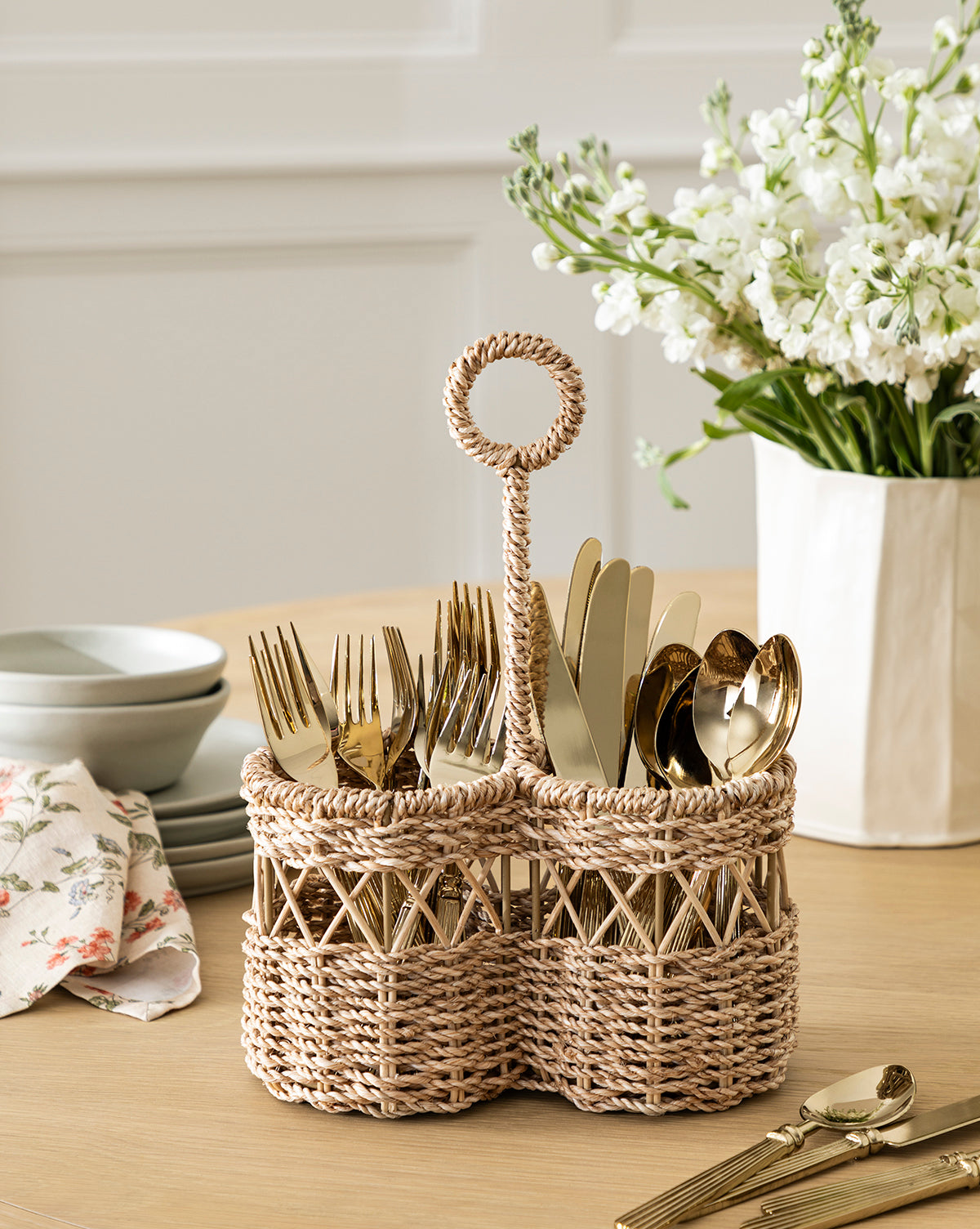 The Stokes Woven Caddy organizes gold-toned forks, knives, and spoons on a wooden table with stacked dishes, a floral napkin, and a white vase of flowers—ideal for adding style to any kitchen counter, by McGee & Co.