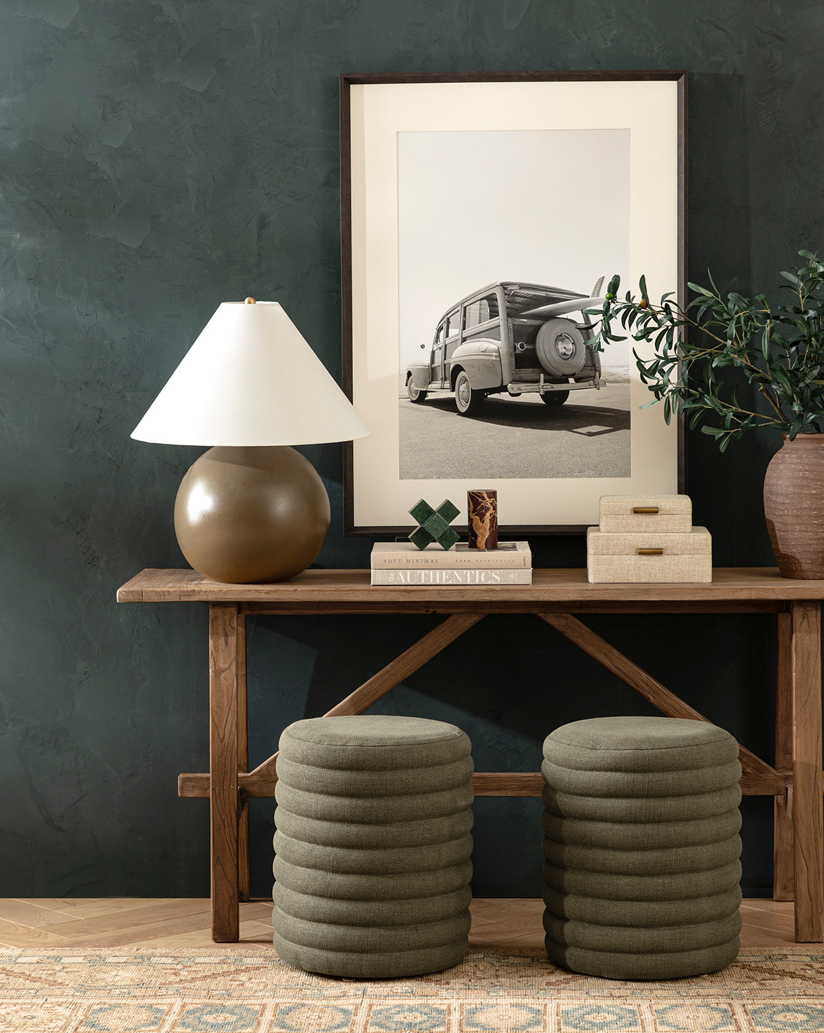 A wooden console table displays the Tess Large Ceramic Table Lamp, books, decorative boxes, and a potted plant. Above hangs a framed black-and-white vintage car photo. Two stacked round green ottomans sit beneath the table, by McGee & Co.