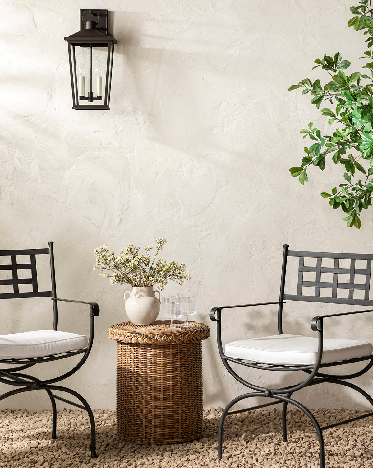 Two black metal chairs with white cushions invite outdoor lounging on a pebbled floor, facing the weather-resistant Haviland Side Table topped with flowers and water. A leafy plant and black wall lantern accent the neutral textured wall, by McGee & Co.