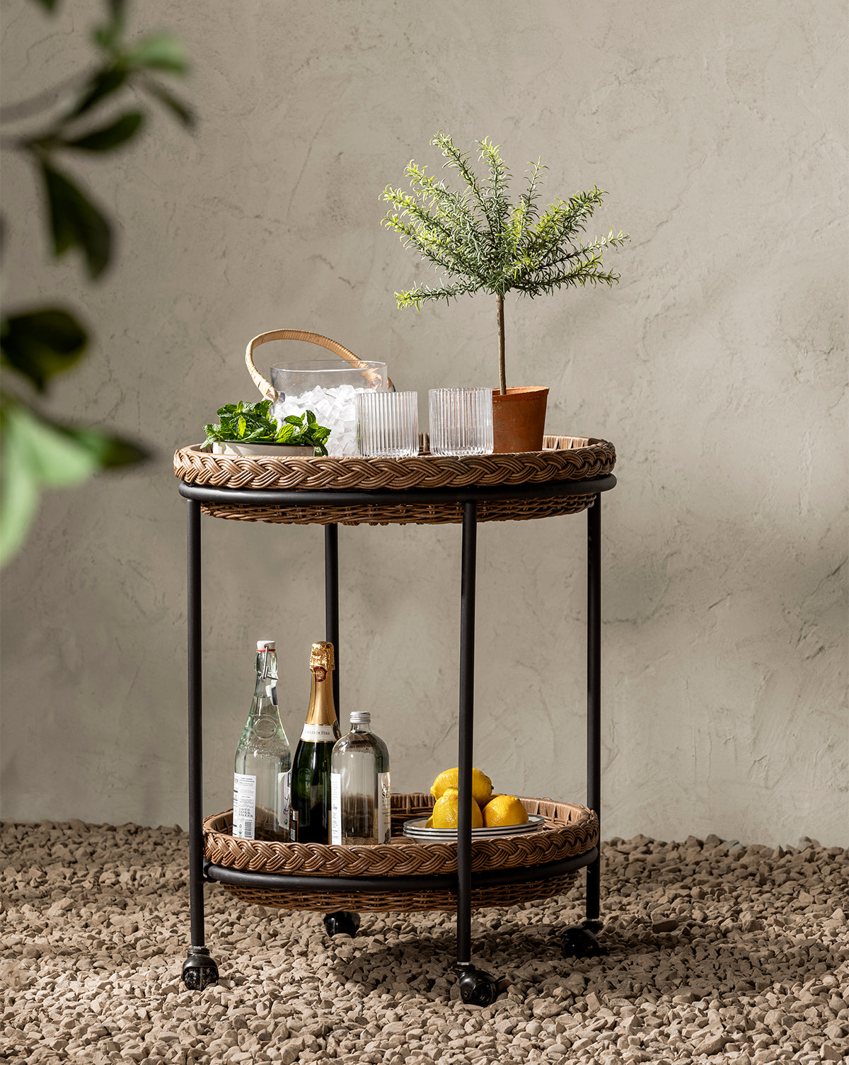 The Haviland Bar Cart, a weather-resistant wicker cart on wheels, sits on pebbles. Its top shelf holds a potted plant, glasses, and an ice basket; the lower shelf displays bottles and lemons—ideal for chic outdoor entertaining. A leafy plant is in front, by McGee & Co.