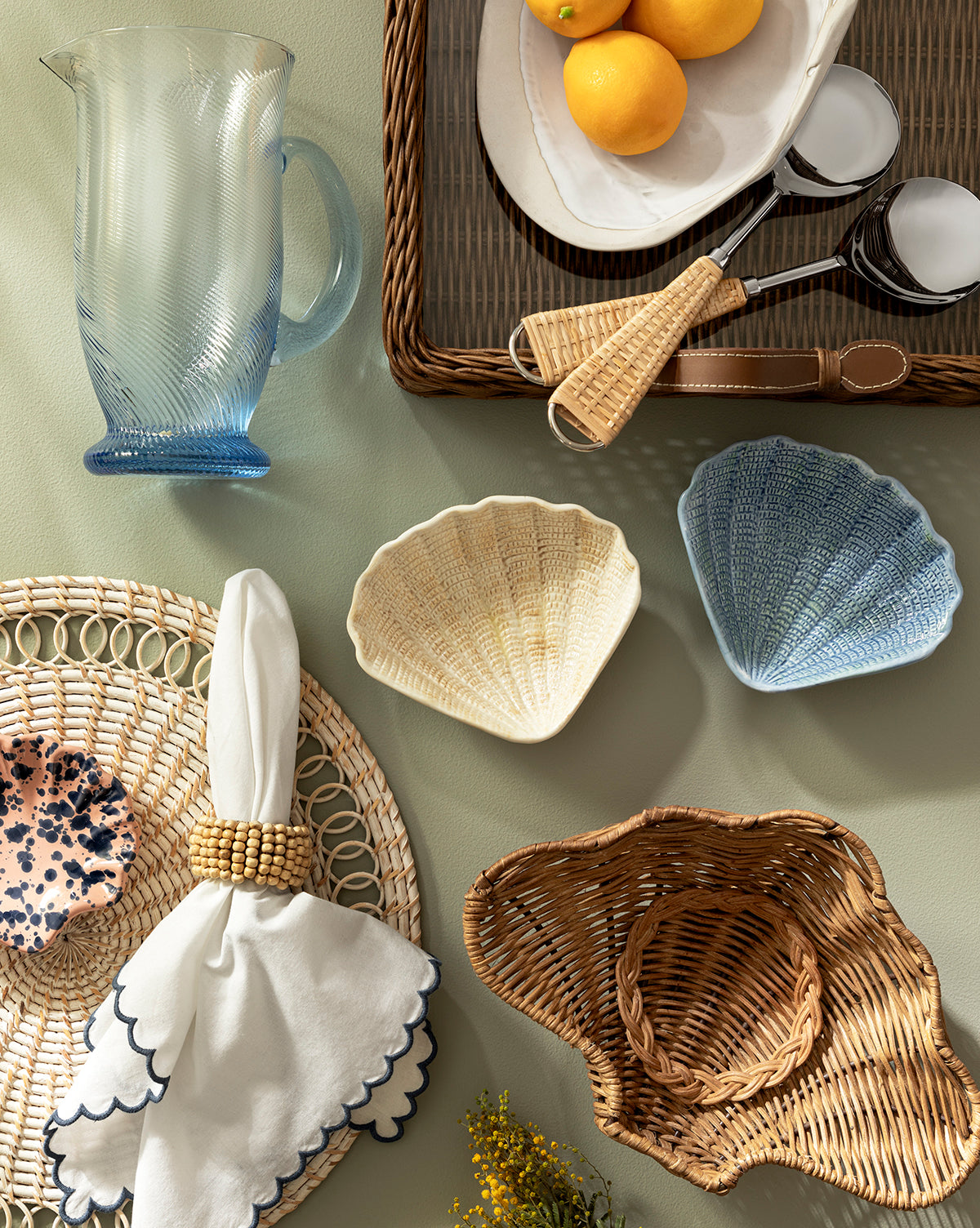A flat lay of coastal-inspired tableware featuring a blue glass pitcher, lemons on a wicker tray, ice scoopers, Aurelie Shell Shaped Bowls (Set of 4), a scalloped napkin on a woven placemat, and a straw sun hat on a light surface, by McGee & Co.