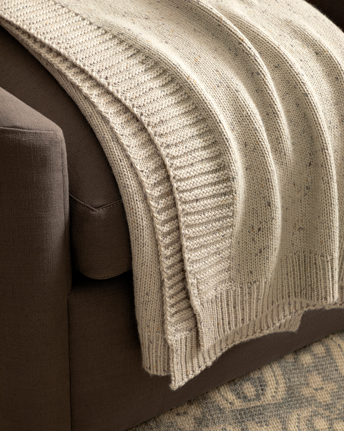 The Makerspalm Otis Knit Throw, a cozy beige wool-blend with ribbed edges, is draped over a dark brown sofa beside a patterned rug, McGee & Co.