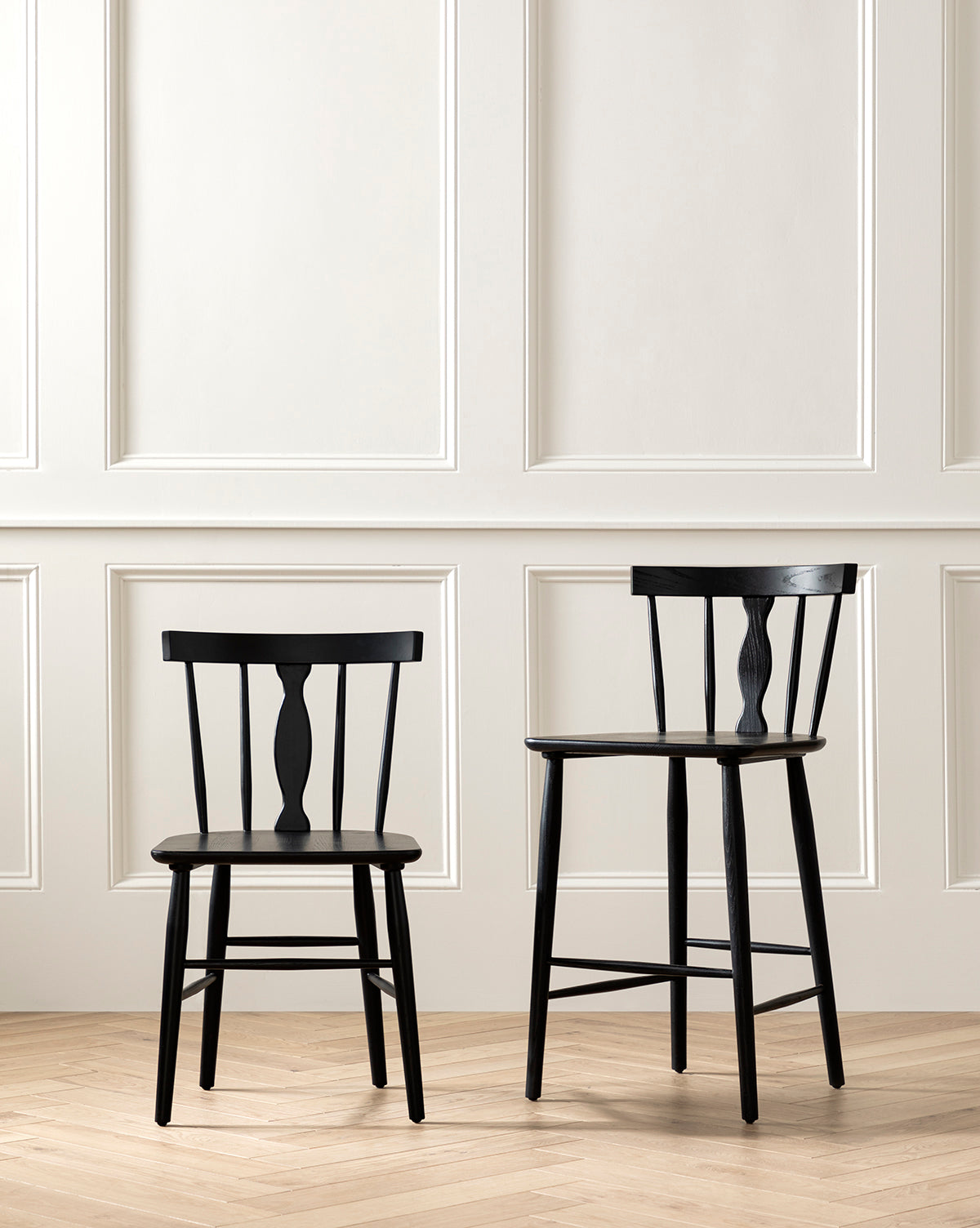 Two black Whittaker Chairs, one shorter and one taller, sit side by side on a light wood floor against a white paneled wall, creating a minimalist dining space by McGee & Co.