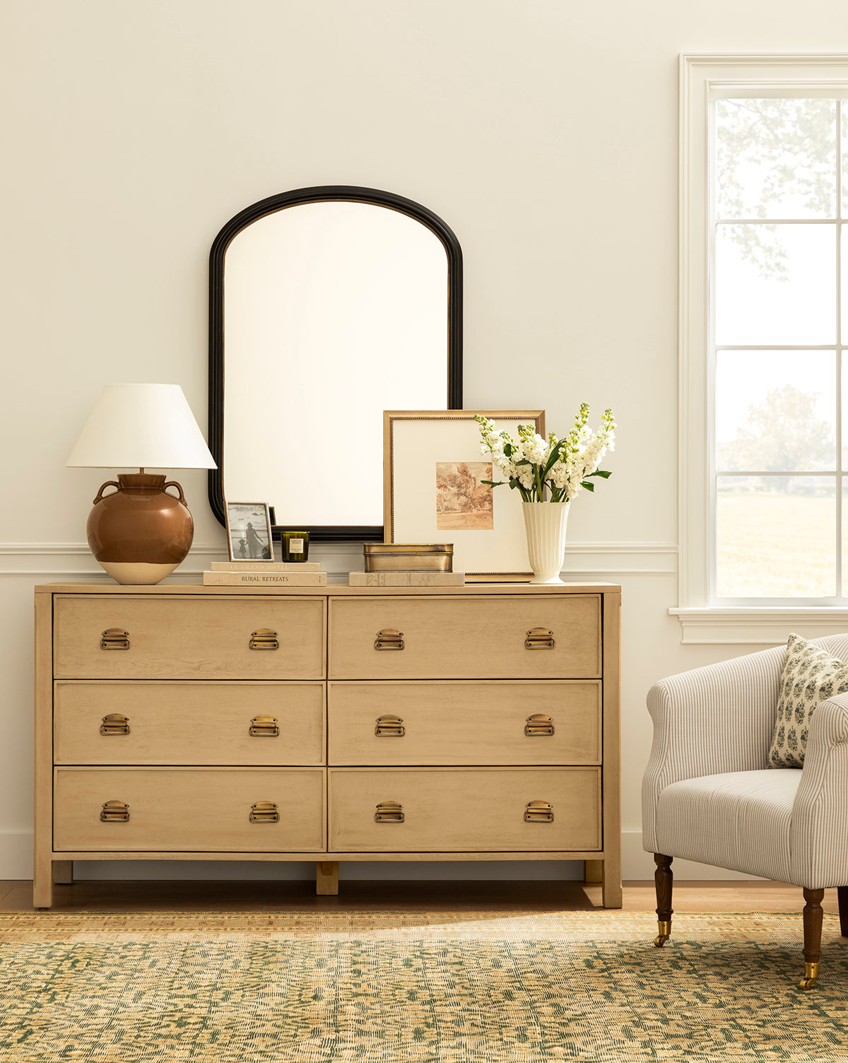 A light wood dresser with gold handles displays a brown lamp, a floral vase, framed art, and books. Above it hangs the Samuel Wall Mirror. To the right are an upholstered armchair and a sunlit window by McGee & Co.