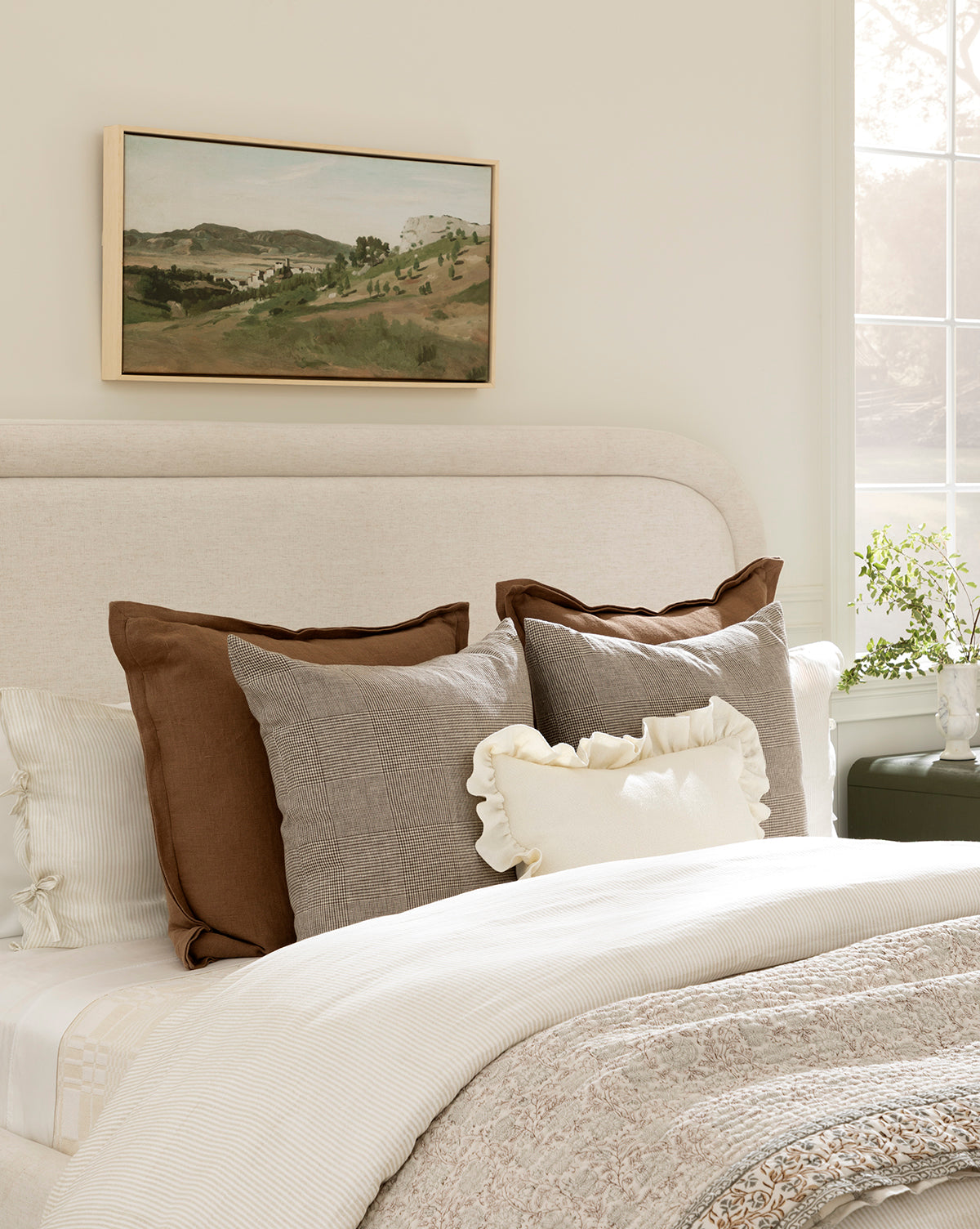 A neatly made bed with cream and brown accents features the Liam Double Flange Pillow Cover, set beneath a landscape painting. Soft light from a window highlights a plant on the nightstand, creating a cozy bedroom atmosphere by McGee & Co.