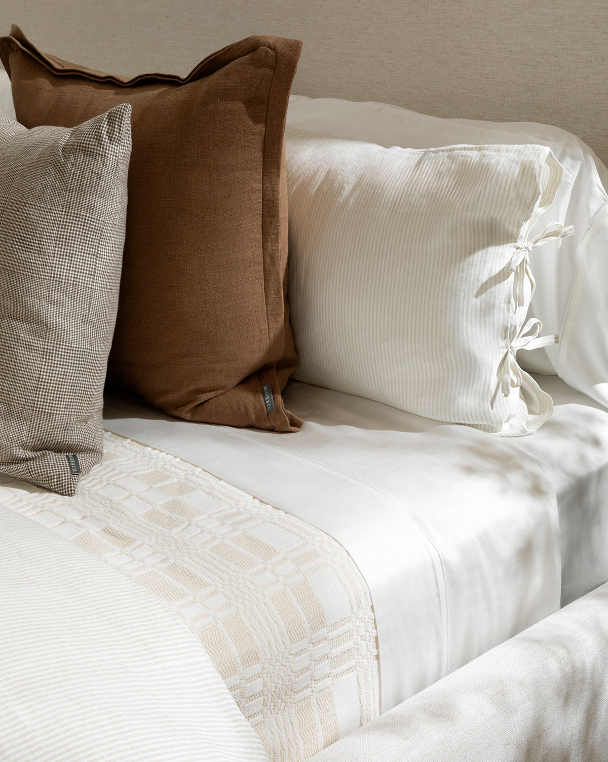 A neatly made bed with crisp white sheets, the Makerspalm Chamberlain Coverlet in beige, two large pillows—one brown, one checkered—and a white pillow with side ties. McGee & Co.