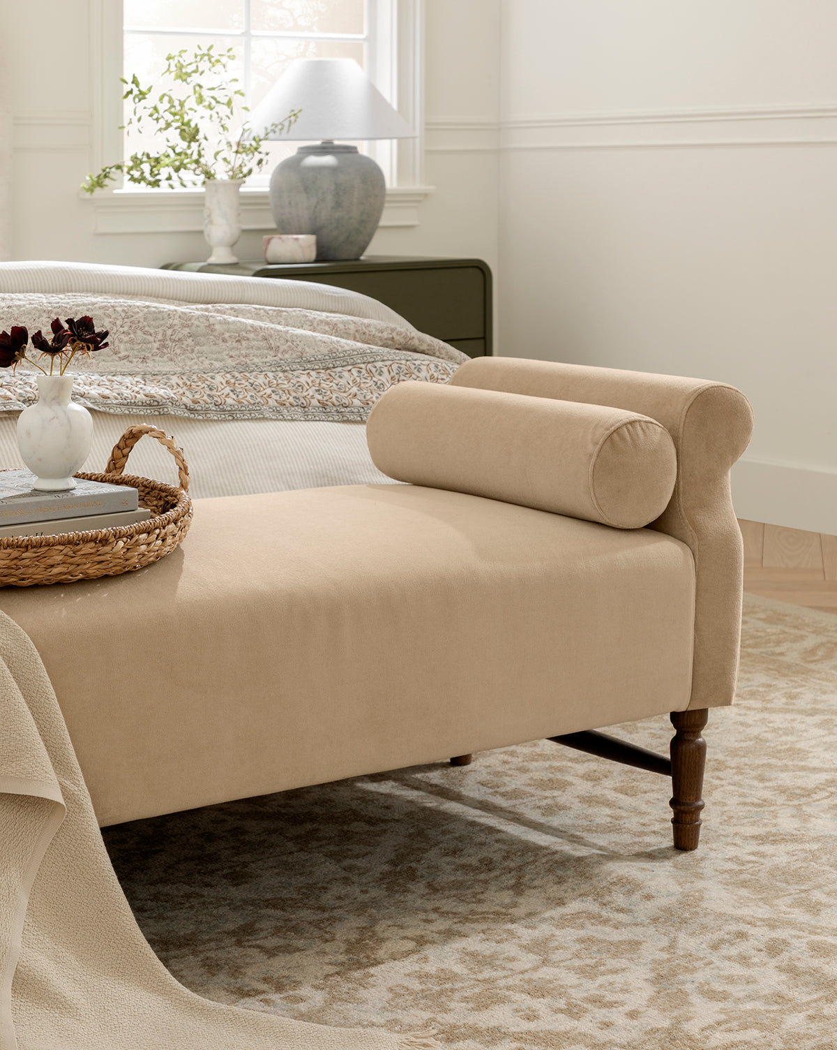 A beige upholstered Asher Bench sits at the foot of a neatly made bed in a cozy bedroom, featuring a cylindrical bolster pillow and topped with a woven tray, vase, and decor. Sunlight pours in from the window behind the bed, by McGee & Co.