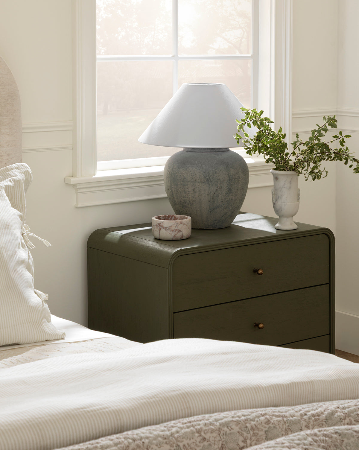 A nightstand holding a Montague Table Lamp, a marble vase with greenery, and a decorative bowl sits beside a neatly made bed with striped bedding near a bright window, by McGee & Co.