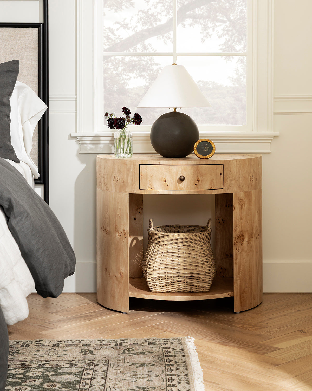 A round wooden nightstand with a drawer and open shelf holds the Boyce Woven Basket below. On top, there’s a lamp, small clock, and a vase with dark flowers, all set by a window next to a bed with gray and white bedding by McGee & Co.