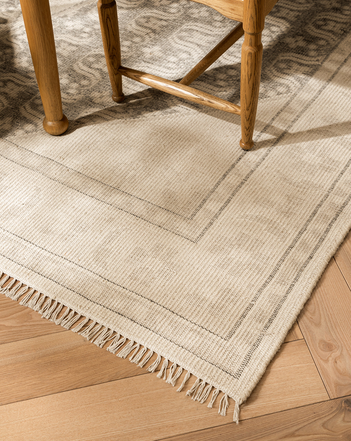 A close-up of the Elin Hand-Knotted Wool Rug in light beige, featuring artisan details, a subtle striped border, and fringed edges on a wooden floor, partially beneath a wooden chair, by McGee & Co.