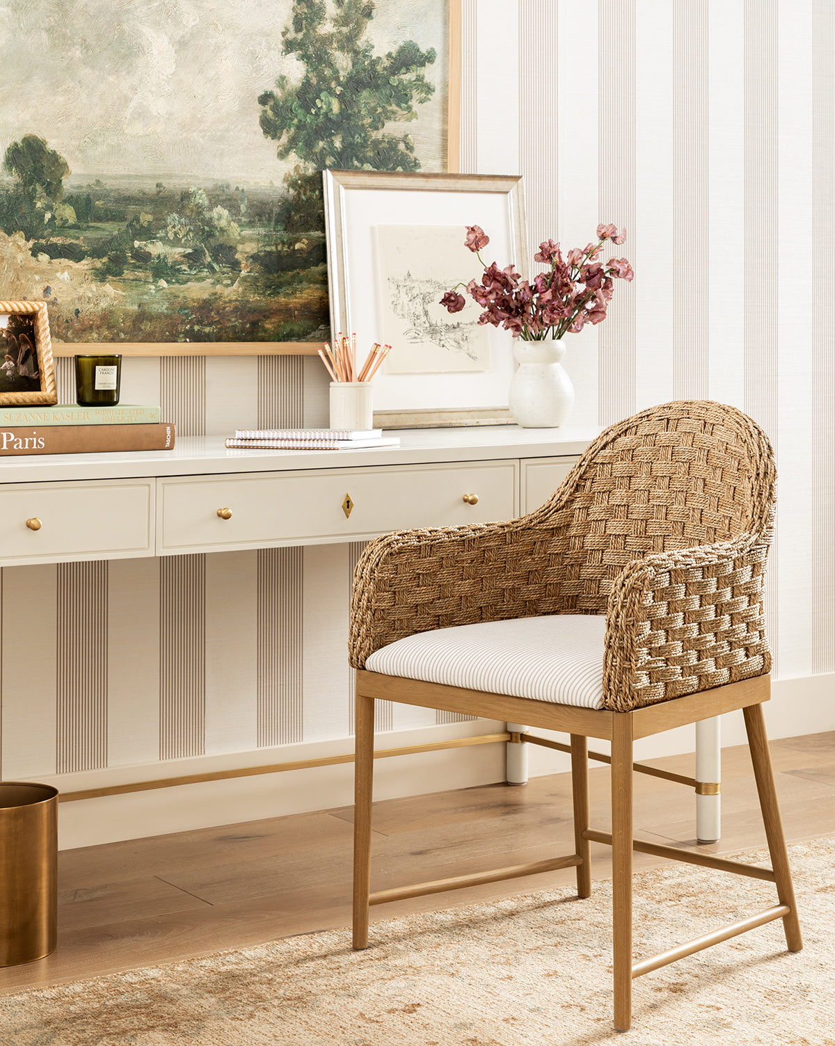 The Molly Arm Chair with a white cushion is placed before a white desk with gold drawer pulls. On the desk are pink flowers in a vase, framed art, books, and pencils. A landscape painting hangs above the setup by McGee & Co.