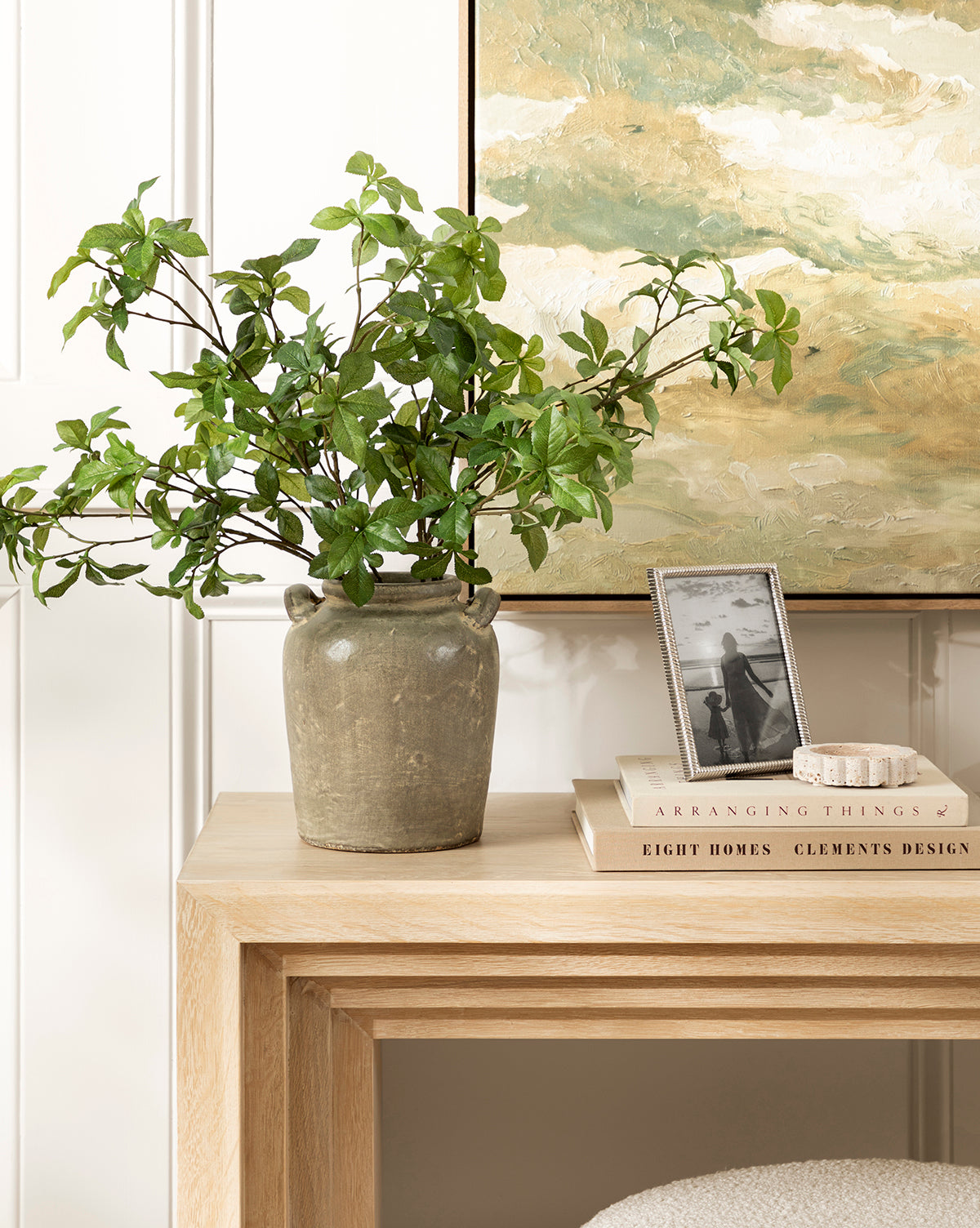A green leafy plant in the Emilio Urn sits on a light wooden console table styled with vintage decor—stacked books, a small dish, and a framed black-and-white photo—beneath a painted landscape, by McGee & Co.