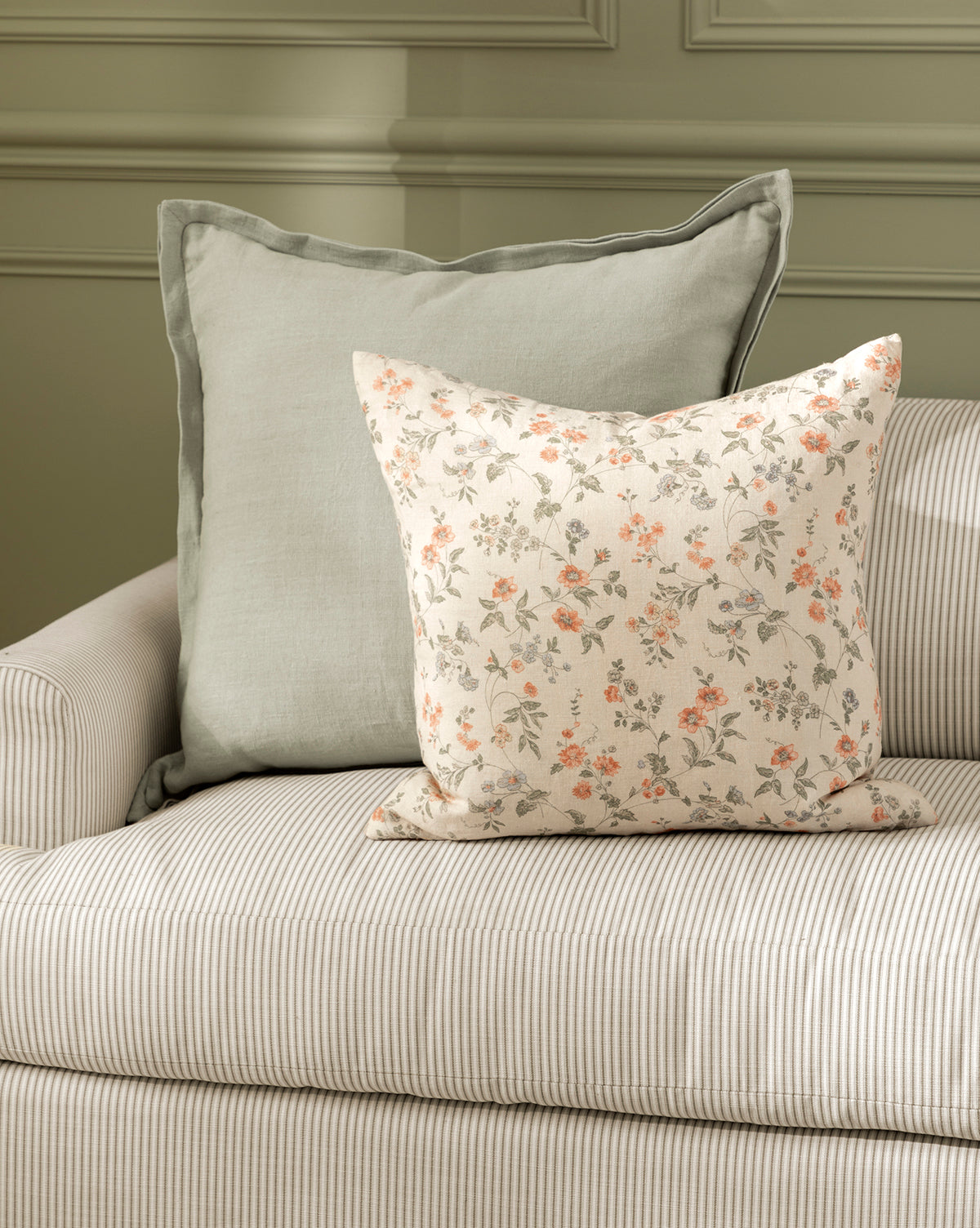 Two Liam Double Flange Pillow Covers—a solid light gray and a cream with orange floral print—decorate a beige striped sofa in front of a pale green paneled wall, by McGee & Co.