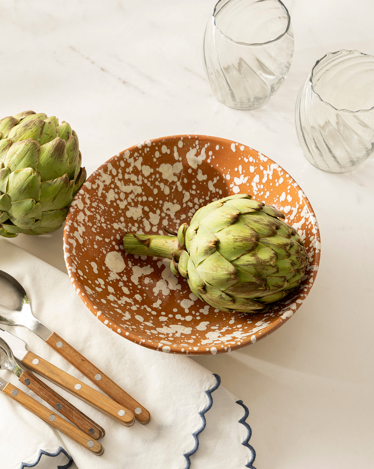 The Splatter Large Bowl, featuring a speckled brown ceramic design and holding an artichoke, sits beside another artichoke, three clear glasses, and wooden-handled utensils on a white napkin with blue trim on a white surface by McGee & Co.