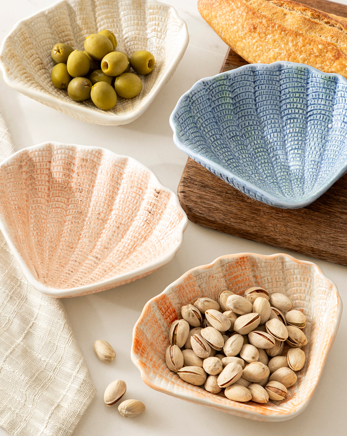 Four Aurelie Shell Shaped Bowls (Set of 4) in pastel tones hold green olives and pistachios. A baguette sits on a wooden board nearby, with a cloth napkin partially visible on the left, by McGee & Co.