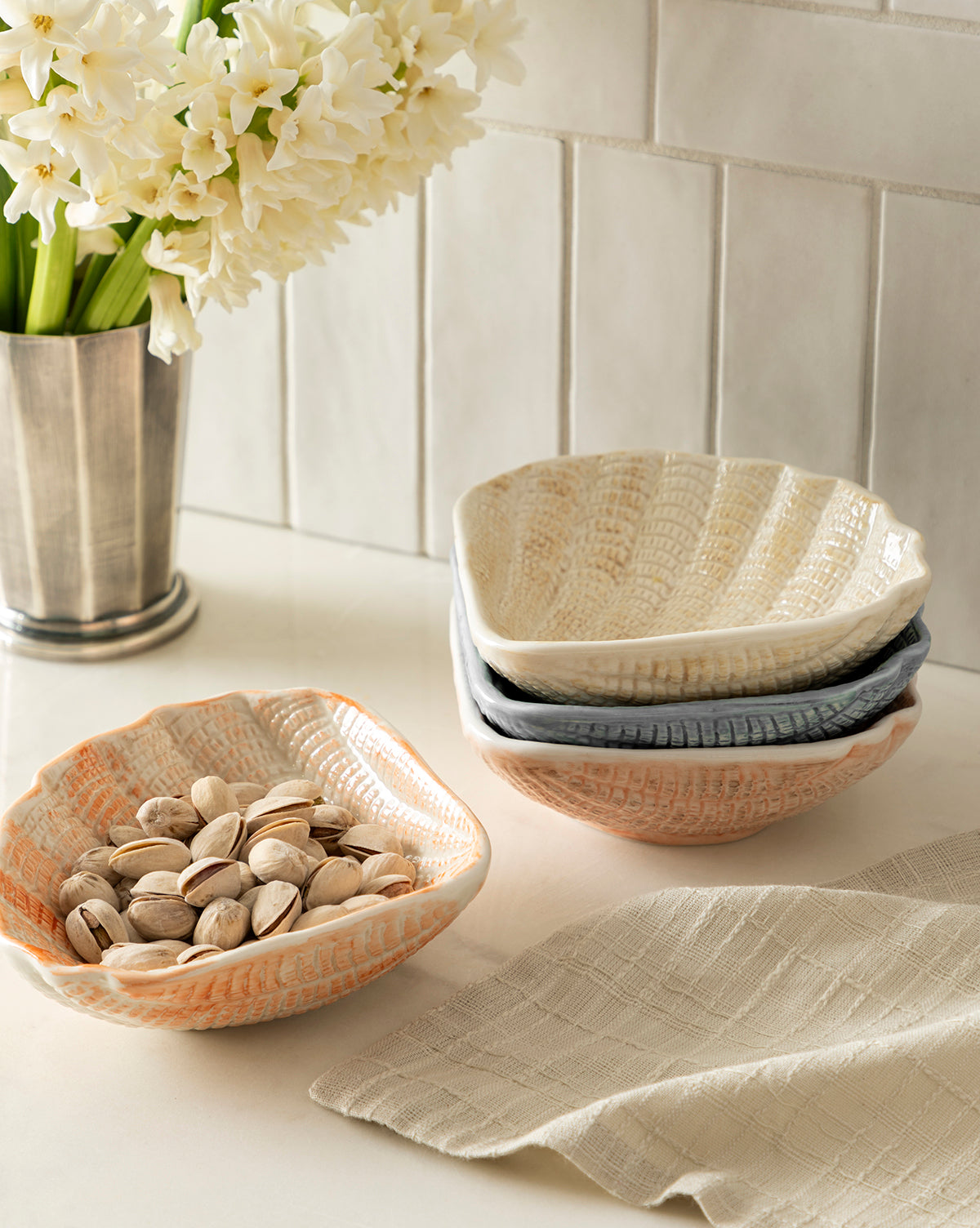 Four coastal-inspired Aurelie Shell Shaped Bowls are displayed on a countertop—one filled with pistachios, two stacked, and one beside them—with a beige cloth and metal vase of white flowers in the background against a tiled wall by McGee & Co.