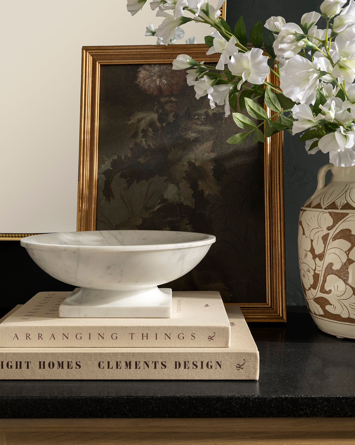 The Tayva Bowl sits atop two stacked design books on a dark countertop, with a framed floral painting and a ceramic vase of white flowers behind it to complete the charming display, by McGee & Co.